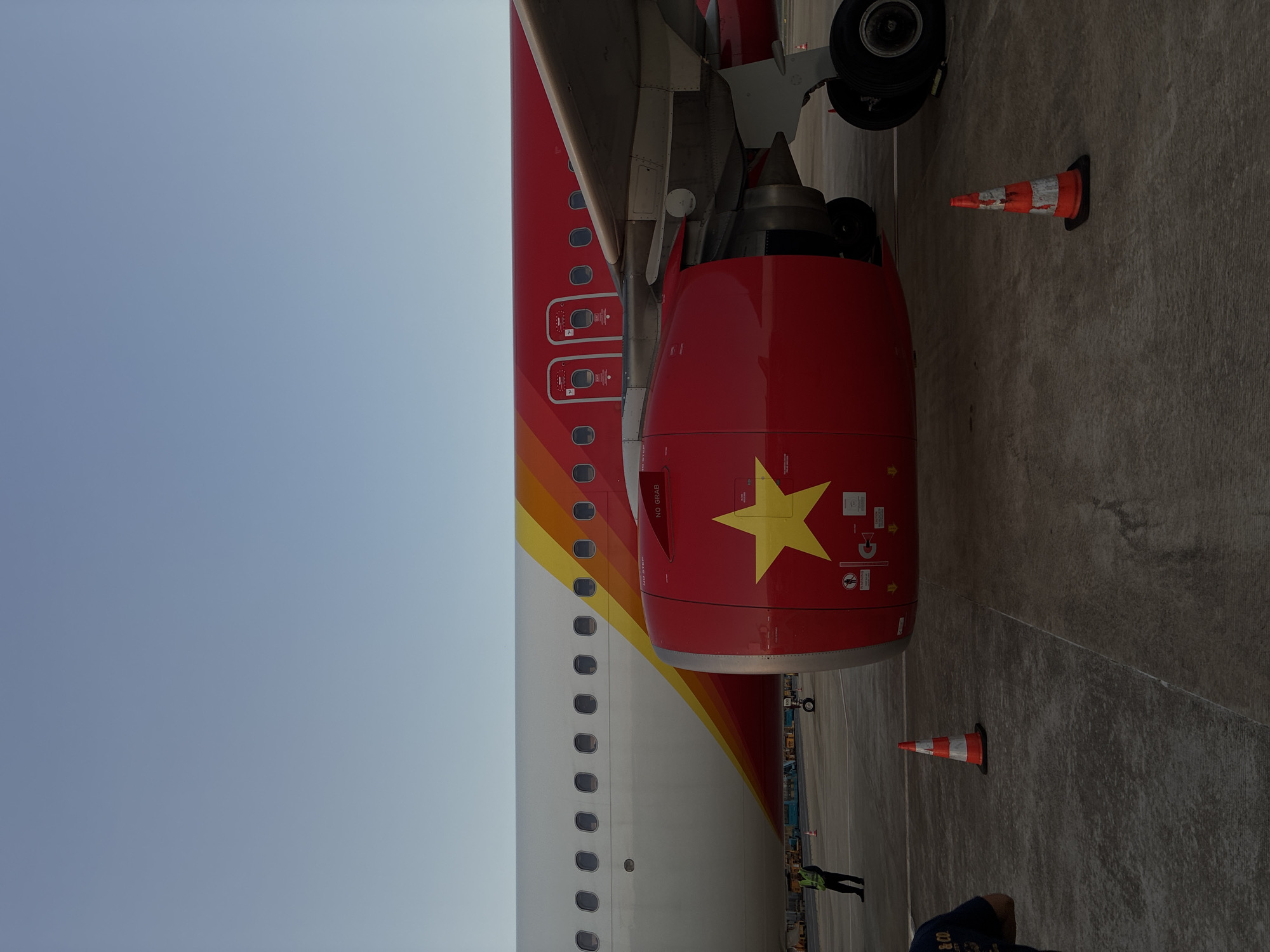 An airplane engine, painted bright red with a yellow star, is prominently featured in the foreground of this image. The airplane's fuselage, white with a red and yellow stripe, extends behind the engine. The scene appears to be on an airport tarmac, possibly at Da Nang International Airport, with a clear blue sky overhead. Several orange and white traffic cones are scattered on the ground, suggesting ongoing ground operations or maintenance. A person in a green vest, likely an airport worker, stands near the wing of the aircraft, and another person is partially visible in the lower left corner. The overall impression is that of an airport environment with an aircraft ready for departure or having just arrived.