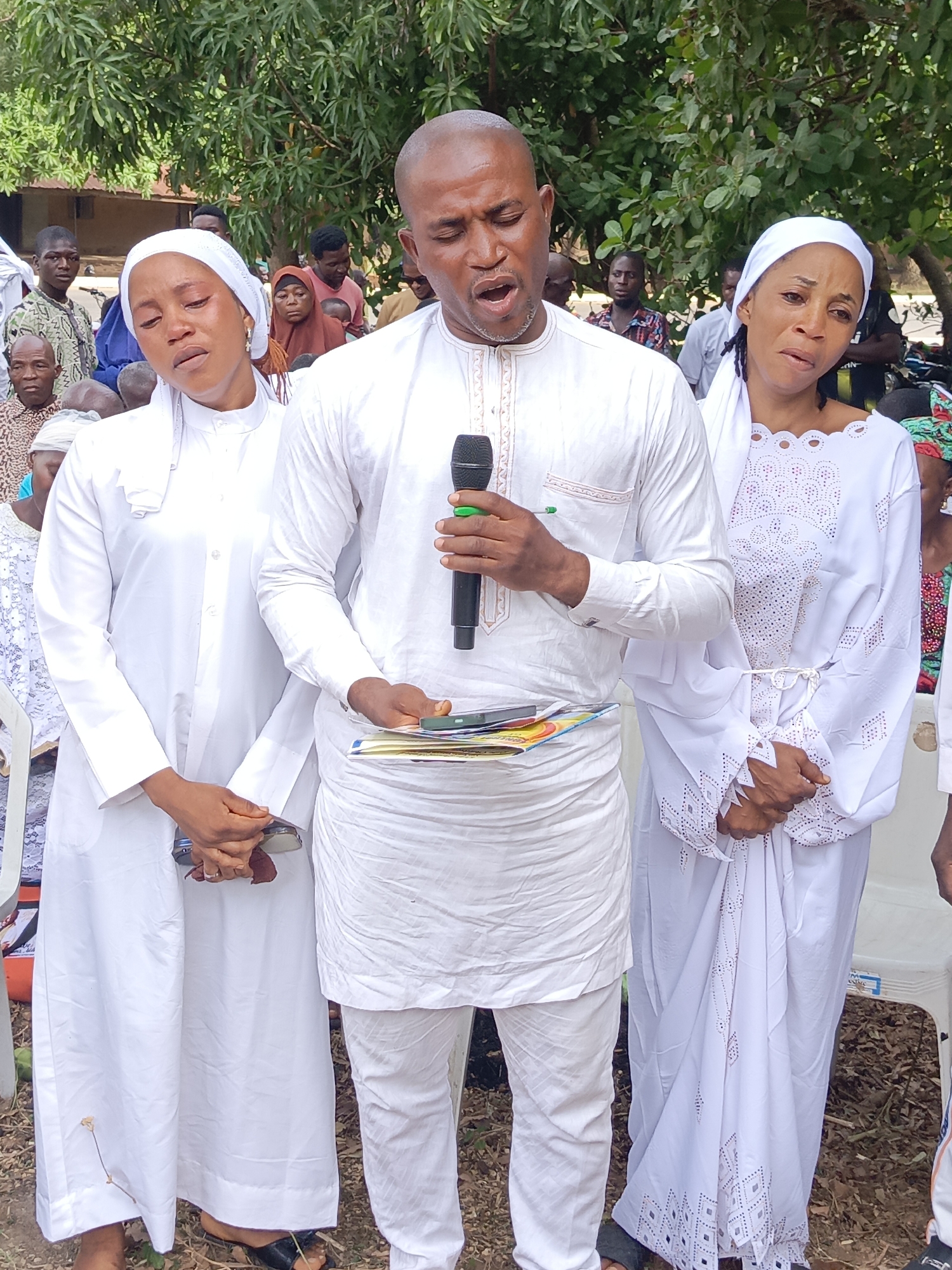 This image depicts an outdoor community gathering in Andaha, Nigeria, centered around three individuals dressed predominantly in white. The central figure is a man wearing a white embroidered tunic and trousers. He is holding a black microphone in his right hand and a stack of colorful books or pamphlets in his left, speaking or singing with his mouth open.
He is flanked by two women, both adorned in white attire and white head coverings. The woman on his left wears a plain white long dress and a simple headscarf. Her eyes are closed or cast downwards, and her face shows deep emotion, possibly sorrow or prayerfulness, with visible moisture around her eyes. She holds a small object, likely a phone, clasped in her hands.
The woman on his right is dressed in an ornate white gown featuring intricate cut-out designs and embroidery, paired with a white headscarf. Her expression also conveys strong emotion, with her mouth slightly open and her gaze directed upwards or away, suggesting distress or spiritual intensity. Her hands are clasped together in front of her.
In the background, a crowd of people, including men, women, and children, are gathered, some standing and others seated on white plastic chairs. The scene is set outdoors amidst green foliage and trees, with a building partially visible in the distance. The bright lighting indicates it is daytime. The overall mood appears solemn and emotionally charged, characteristic of a religious or communal ceremony. No discernible text is visible.
