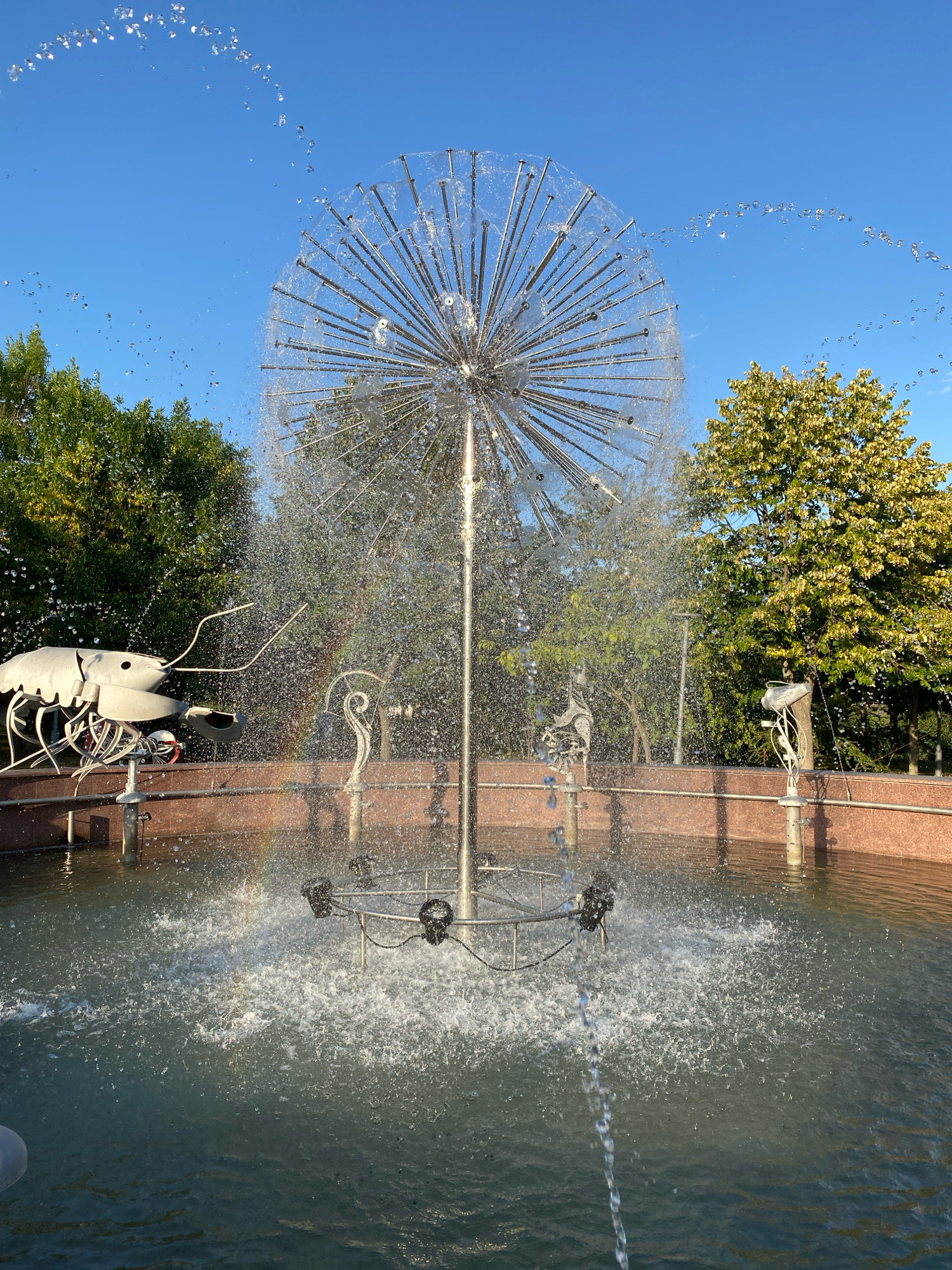 The image depicts a dandelion-shaped fountain spraying water, set against a backdrop of trees and a clear blue sky. Located in Chișinău, Moldova, specifically in the central park area near the President's Administration Building, this fountain is a popular landmark. The fountain's metallic structure, resembling a sphere with radiating rods, is the central focus. Water streams out from various points, creating a dynamic display. Two decorative metallic sculptures, one resembling a shrimp and another a stylized bird, are positioned on the edge of the fountain basin. The basin itself is lined with red granite. Sunlight illuminates the scene, casting subtle reflections on the water's surface and creating a rainbow effect from the spray. The surrounding environment suggests a well-maintained park, with lush green trees and a clear sky indicating fair weather. No people are visible in the image.