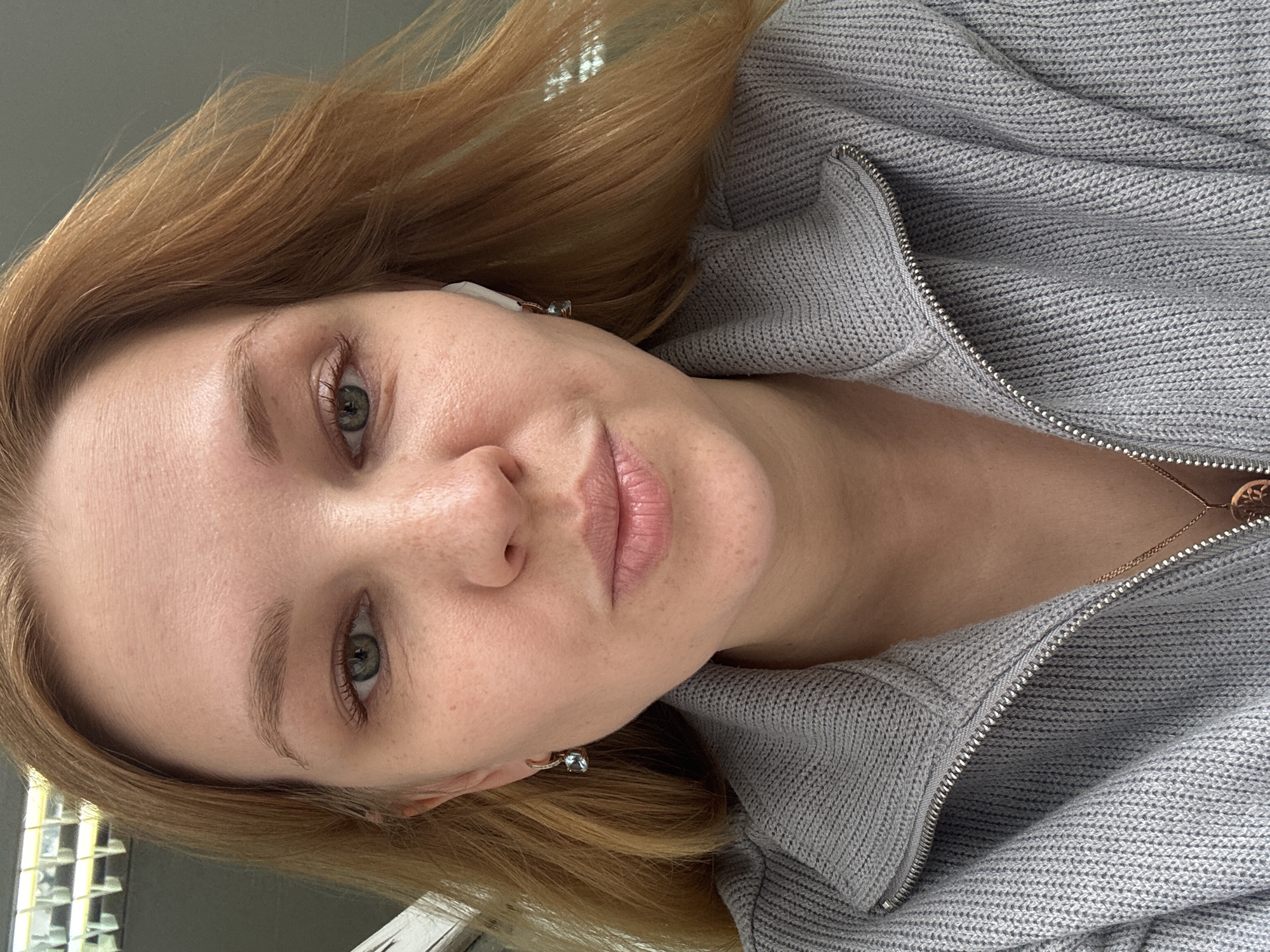 A close-up portrait shows a fair-skinned woman with red-blonde hair and green-grey eyes. She has freckles on her face. She is wearing a light grey zip-up knit sweater and a delicate gold necklace. A small earring is visible in her left ear, and an earbud is in her right. No location is specified.