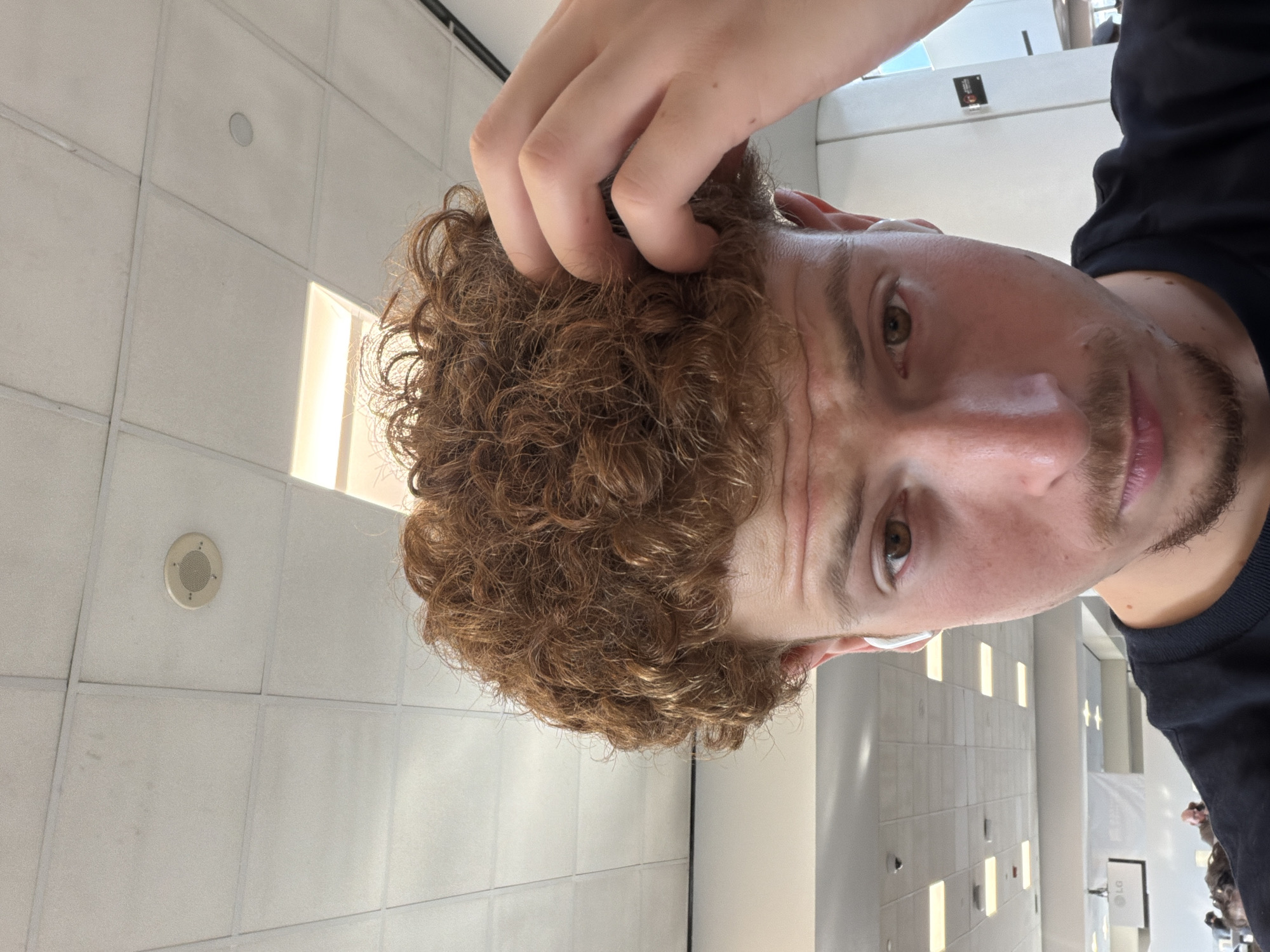 A person with brown, curly hair and a light complexion is looking at the camera. The person has brown eyes, a goatee, and is wearing a dark-colored shirt. The person's hand is touching their hair. The background shows a tiled ceiling with recessed lighting. In the distance, there are glimpses of a brightly lit corridor with retail displays. Considering the architecture, lighting, and layout, this image appears to have been taken inside Newark Liberty International Airport (EWR) in Newark, New Jersey.