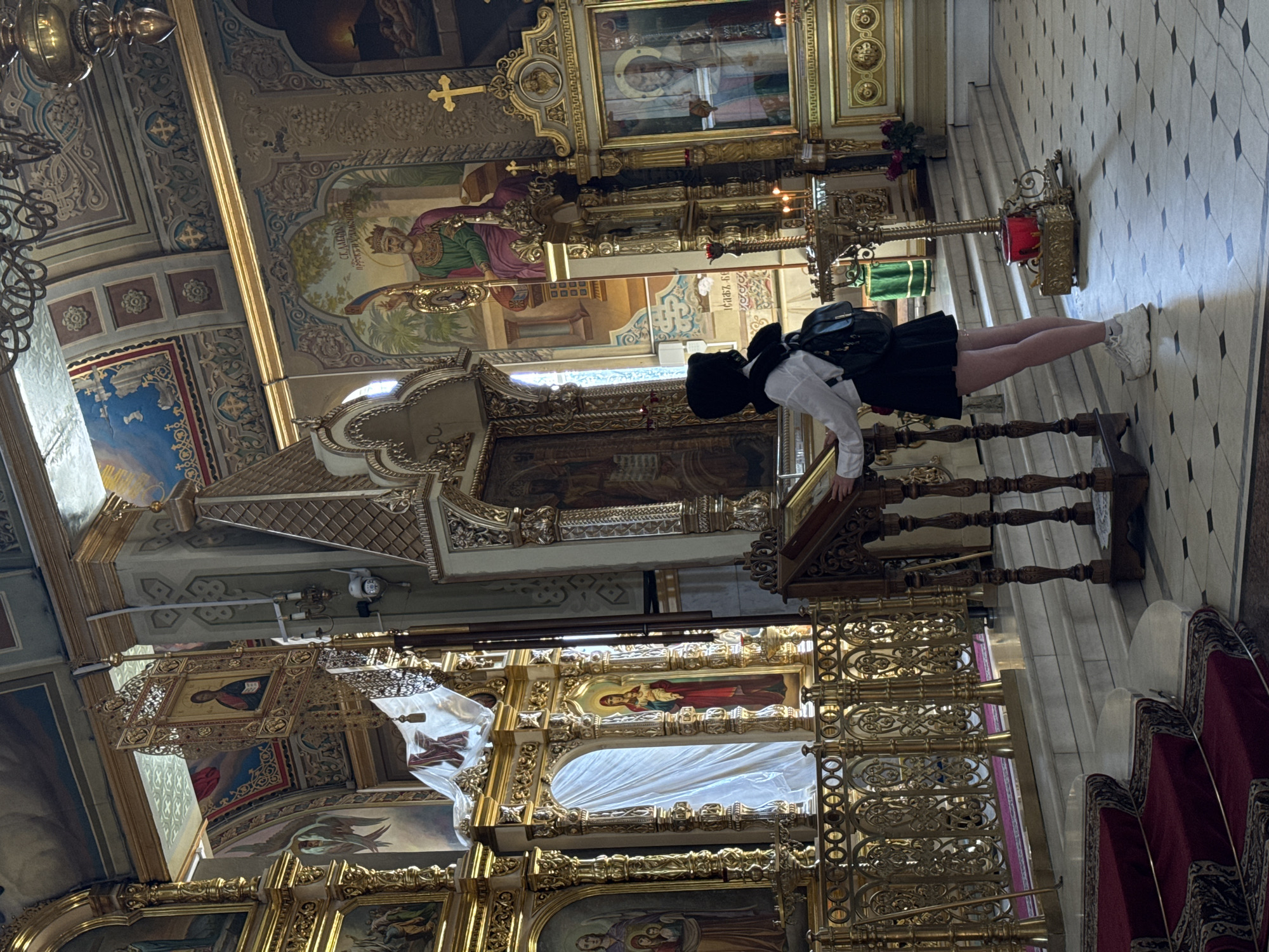 In the interior of a church, a young person wearing a black head covering, a white collared shirt, and a black pleated skirt stands at a wooden lectern, facing away from the camera. They have a black backpack on and are wearing white sneakers. The church features ornate gold detailing on the iconostasis, painted religious murals on the walls and ceiling, and marble flooring with a black tile grid pattern. Red carpeted stairs are visible in the foreground. Several icons are displayed within gilded frames, and candles are lit in candelabra. Sunlight streams in from an unseen source, illuminating parts of the scene. The overall environment is that of a richly decorated religious space.