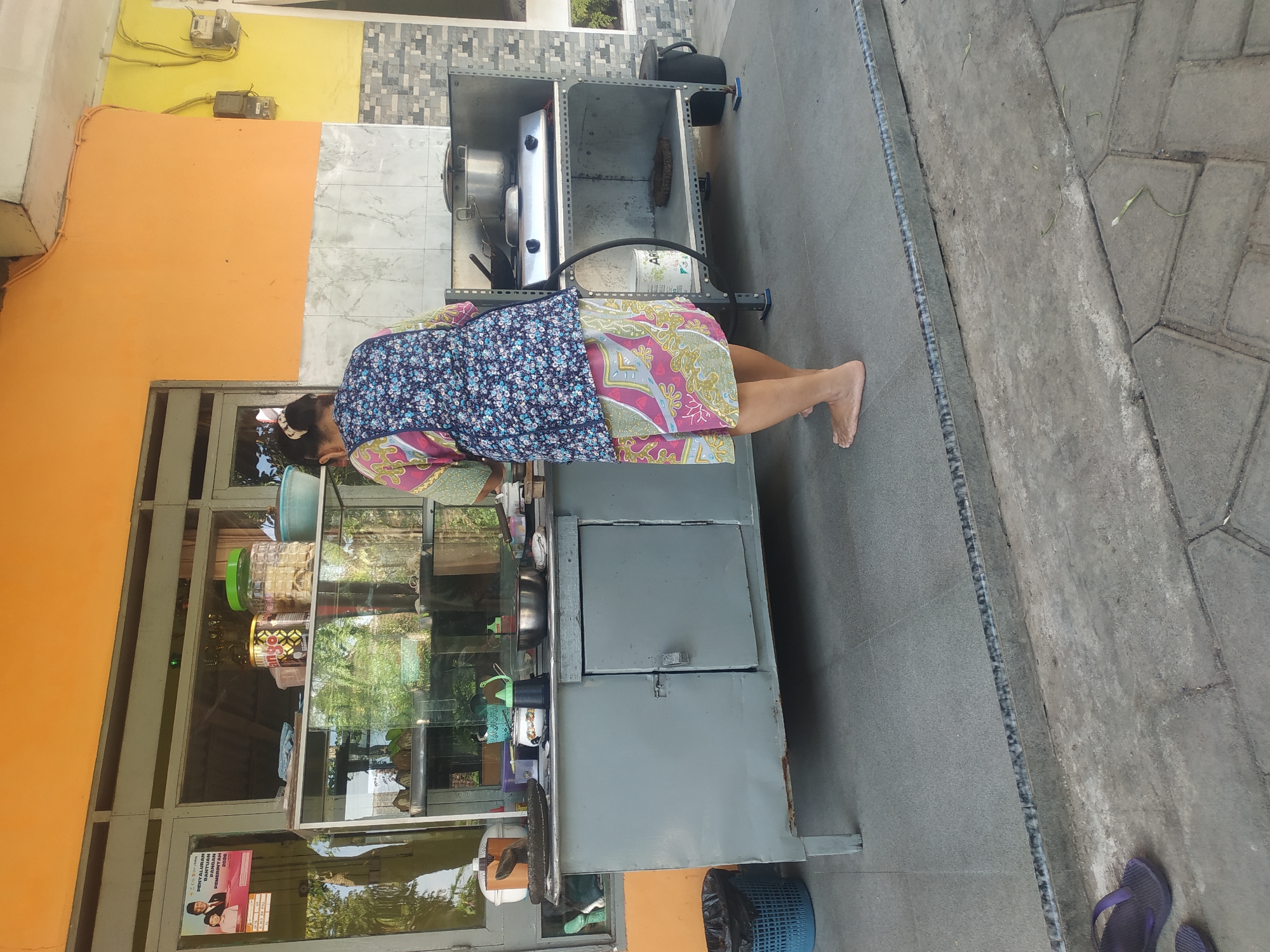 In Jember, Indonesia, an overhead view captures a woman with her back to the camera standing at an outdoor food stall. She is barefoot and wearing a floral print apron over a patterned dress. She appears to be cooking or preparing food at a counter. The stall features a glass display case filled with various items, a gas stove with a pot on it, and several containers on shelves. The area around the stall is paved with concrete tiles and there are some plants visible in the background. A blue trash can is located to the left of the stall. The stall is set against a wall painted in bright orange and yellow. A sign with Indonesian text is visible on the glass display.