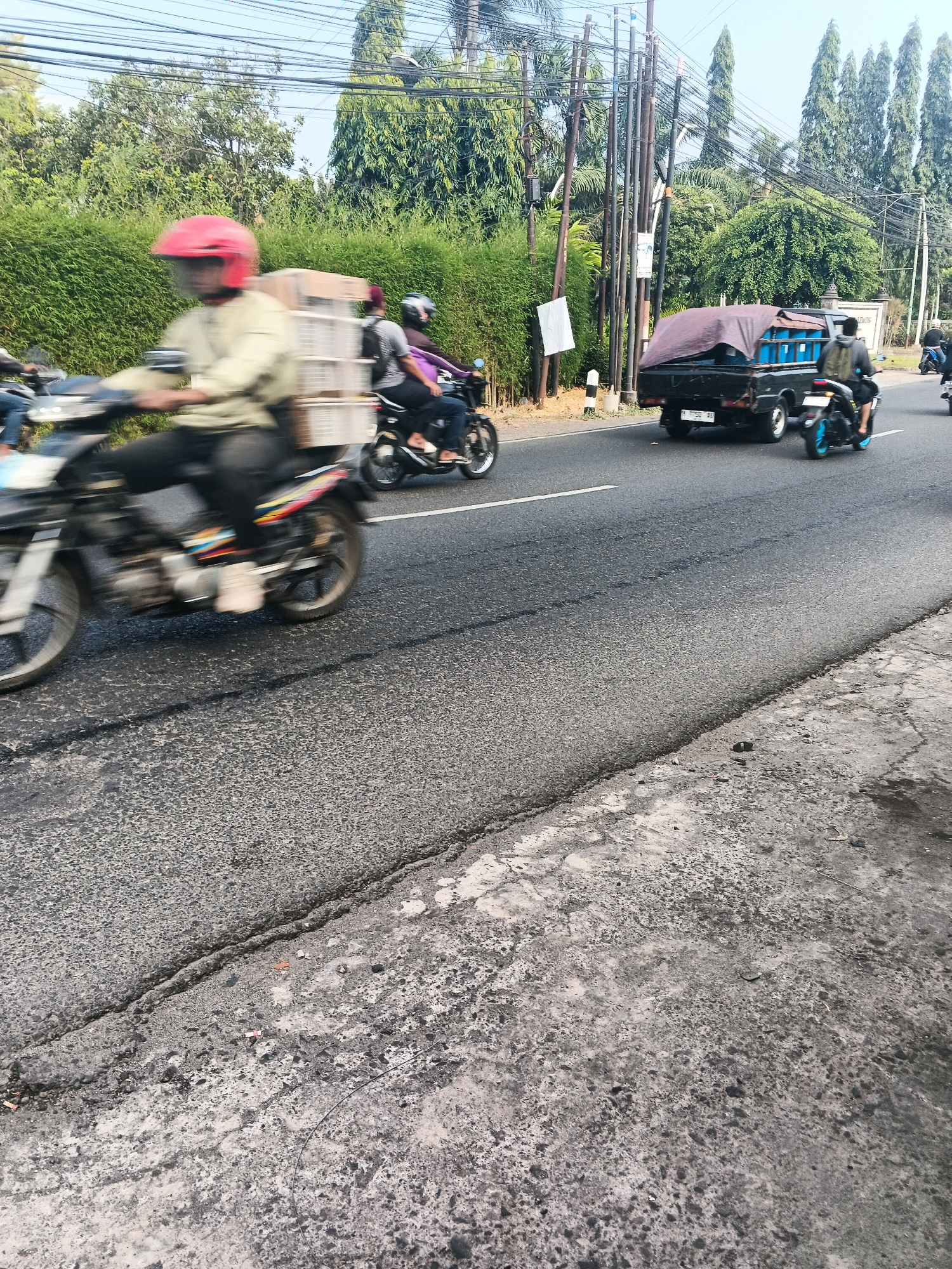 A street scene in Ungaran, Indonesia, captures the blurred motion of vehicles on a paved road. In the foreground, a motorcycle rider in a red helmet and a light-colored jacket is moving from left to right, with a stack of white crates behind them. Another motorcycle rider is partially visible to the left. Further down the road, a black pickup truck with a blue tarp covering its cargo moves in the same direction. To the right of the truck, a motorcycle rider in a black shirt and backpack is also on the road. The environment includes lush green foliage lining the road and utility poles with numerous wires overhead. The road itself appears to be in moderate condition with visible cracks and patches. The sky is clear and bright.