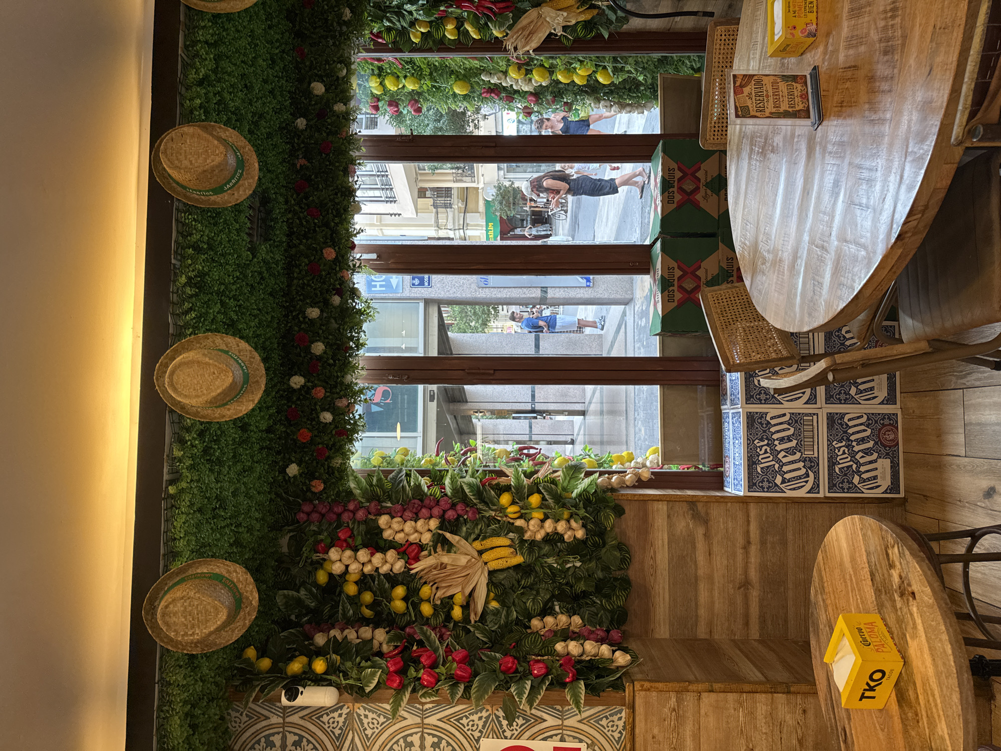 The image depicts the interior of a restaurant or bar in Valencia, Spain, decorated with a vibrant, festive theme. Straw hats adorn a lush green ceiling installation, interspersed with artificial fruits and vegetables like lemons, peppers, garlic, and grapes. Three large windows offer a view of a pedestrian street outside, showing people walking and buildings. Inside, wooden tables and chairs are set up, some with reserved signs. In the foreground, a round wooden table has a yellow napkin dispenser with "TKO Tacos Paloma" branding. Another table features a reserved sign and yellow boxes. Stacks of "Jose Cuervo" and "Dos Equis" boxes are visible near the windows, suggesting a Mexican or Spanish theme. The overall ambiance is casual and lively, likely a popular spot for dining and drinks in Valencia.