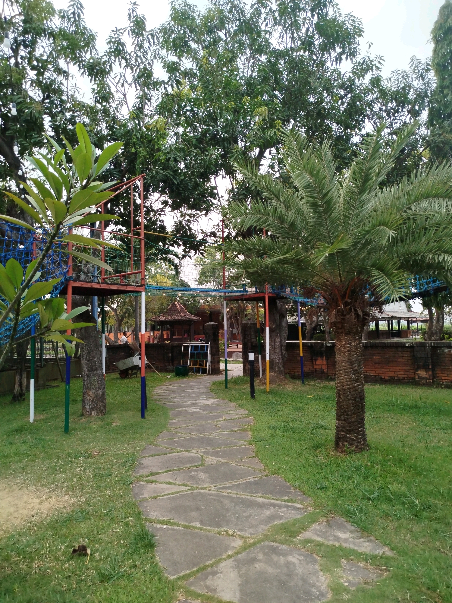 The image depicts an outdoor scene in Bugangin, Indonesia, featuring a winding stone pathway through a grassy area. To the left of the path, a multi-level play structure with red and blue elements is visible, partially obscured by lush green foliage from a nearby plant. A date palm tree stands prominently on the right side of the path. In the background, a brick wall separates the grassy area from a building with a distinctive roof, suggesting a park or recreational facility. The sky above is overcast, with the tops of several large trees framing the scene. The overall environment appears to be a well-maintained public space.