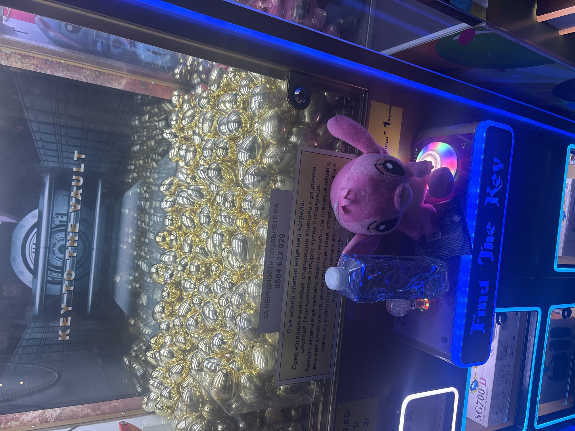 This image captures a brightly lit scene, likely indoors, featuring a "Key To The Vault" prize machine. The machine is filled with gold-colored, egg-shaped capsules. A pink stuffed animal, resembling the character "Angel" from the Disney movie "Lilo & Stitch," sits atop the machine, next to a partially visible blue water bottle. The machine features a blue neon sign that reads "Find The Key." There is a sign displayed on the glass near the capsule eggs in Bulgarian, which includes a phone number. The phone number in the sign (0884 622 929) allows us to place the photograph in Bulgaria. Based on the phone number, it is highly likely the image was taken in Bulgaria, but not explicitly the city of Plovdiv. However, the text on the glass indicates the presence of prizes within the eggs, specifically mentioning colored keys that can open boxes for gifts. This suggests the location is an arcade or amusement center. The image also contains visible text indicating the cost of a game (1 жетон, or 1 token), and other pricing information in Euros.