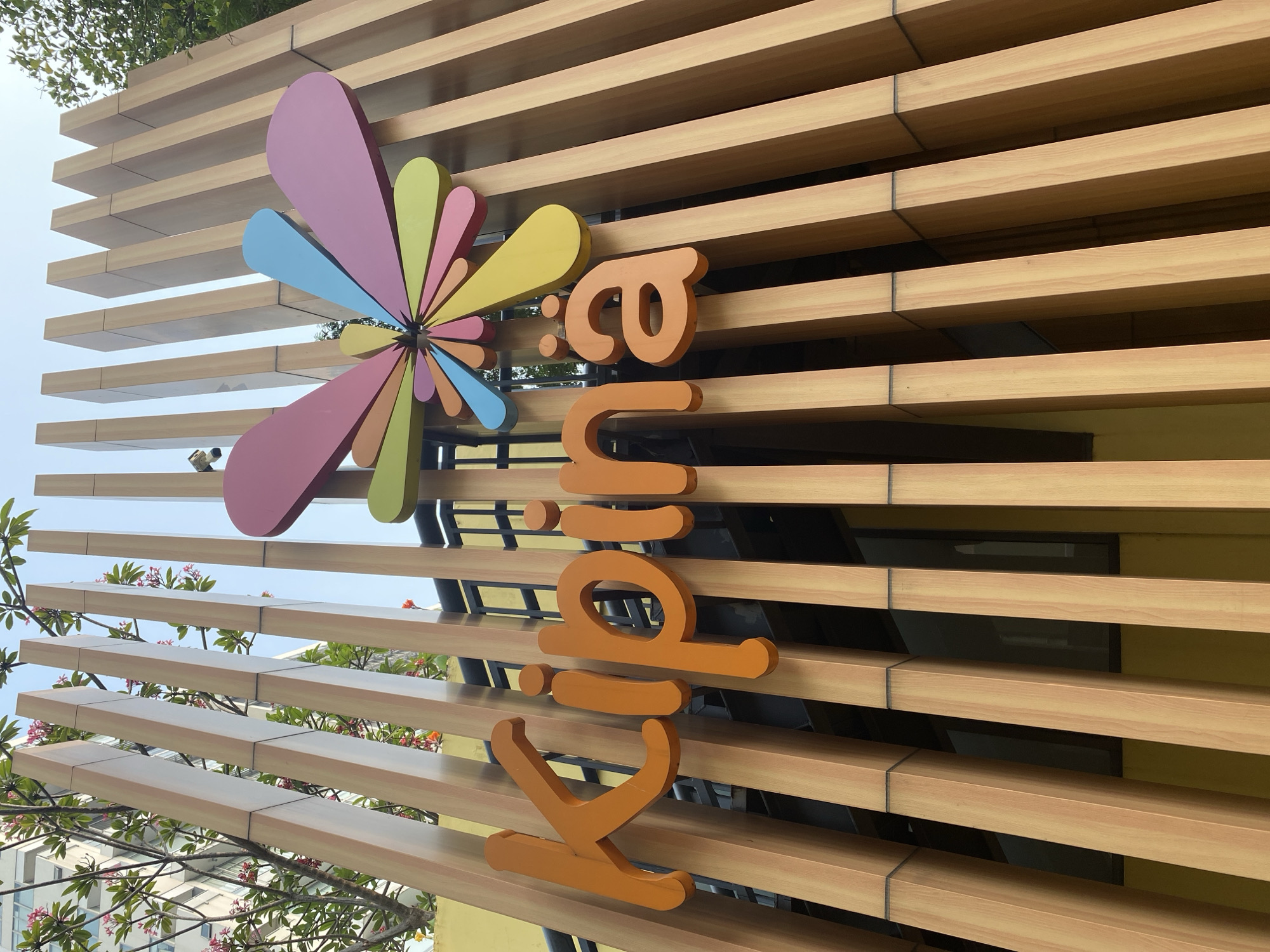 The image shows the exterior signage of "Kipina" in North Jakarta, Indonesia. The sign consists of large, orange, three-dimensional letters spelling out "Kipina". Above the text, a colorful, abstract floral design is mounted. The signage is set against a backdrop of vertical wooden slats and a bright blue sky. Some greenery and parts of a building are visible in the background. The perspective is from below, looking up at the signage.