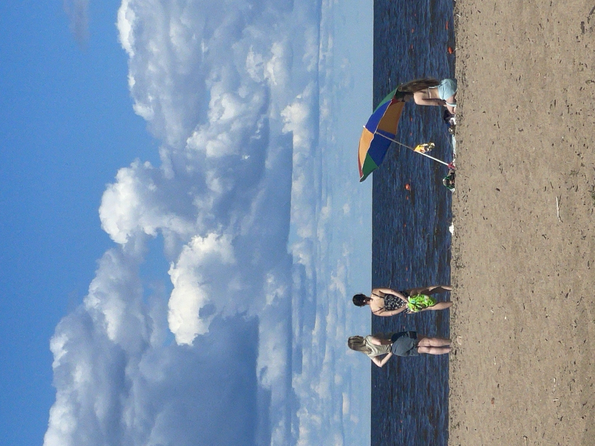 The image depicts a scenic view of a beach in Belozersk, Russia, on a sunny day with a bright blue sky and scattered white clouds. The foreground is a sandy beach, and the background features a large body of water, likely Lake Beloye, which is characteristic of Belozersk's location.
In the mid-ground, three individuals are present. Two women stand near the water's edge, facing away from the camera. The woman on the left wears a light-colored sleeveless top and dark shorts. The woman to her right is in a patterned one-piece swimsuit and has a bright green sarong draped over her arm. Further to the right, under a colorful beach umbrella, a third person, appearing to be a woman, sits on the sand, also facing the water. She is wearing a light-colored bikini.
The umbrella is tilted, casting a shadow. Various items, possibly beach bags or towels, are scattered on the sand near the umbrella. The water appears dark and calm, with a few distant orange buoys visible. The overall atmosphere suggests a leisurely day at the beach.