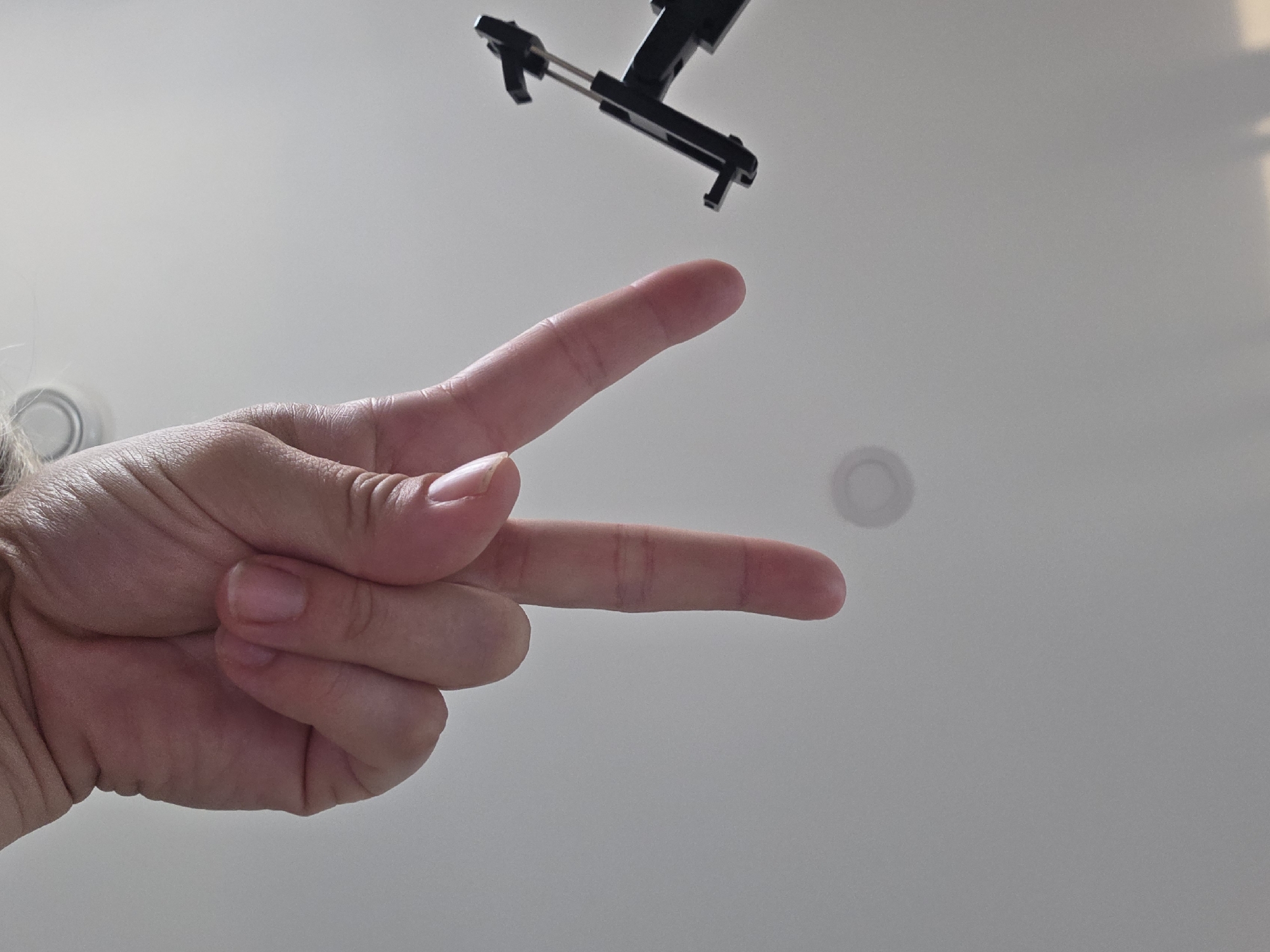 An overhead, low-angle shot shows a person's hand making a peace sign against a plain, light-colored ceiling. A black object, possibly a phone mount or tripod attachment, is visible in the upper left corner of the frame. The ceiling has a circular indentation or light fixture in the upper middle portion of the image. The lighting is diffuse and appears to be from an overhead source. The scene provides no information to determine its location in the city of Grünstadt, Germany.