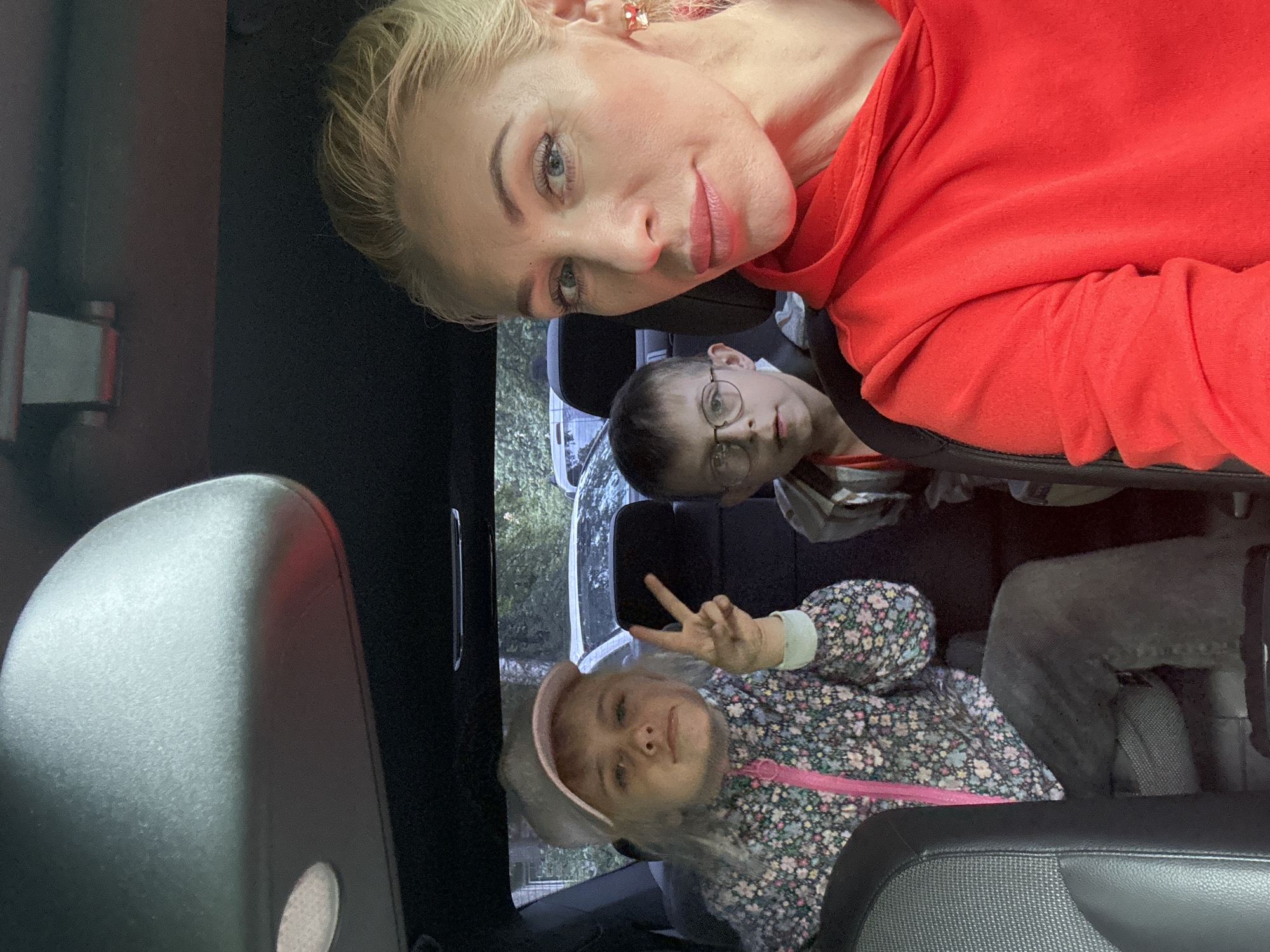 An overhead shot captures a woman and two children in the back seat of a car, seemingly on a road trip or outing. The woman, positioned in the foreground, leans into the frame, her blond hair pulled back, and she wears a bright red hoodie. Her expression is neutral as she looks directly at the camera. To her left, a young girl with blond hair, wearing a floral jacket and a pink baseball cap, faces forward and makes a peace sign with her right hand. Beside her, a boy with short brown hair and glasses is also seated. He looks to the left of the frame with a neutral expression. The interior of the car is visible, with dark upholstery and seatbelts. Outside, through the car's windows, glimpses of trees and other vehicles suggest an outdoor setting, possibly in a suburban area or park. The lighting suggests it is daytime. The overall scene appears candid, capturing a moment during a family outing. There is no explicit indication of the location being Saint Petersburg, Russia.