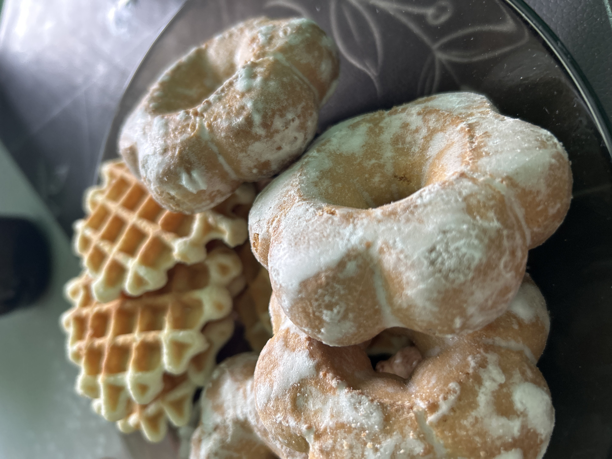 In a close-up shot, a pile of baked goods sits on a dark, possibly glass, surface. Dominating the foreground are several ring-shaped cookies, generously dusted with white powdered sugar, which creates a marbled effect against their golden-brown surface. These cookies appear to have a slightly irregular, perhaps homemade, texture. Nestled behind and to the left of the cookies are several stacked waffle cookies. These waffles are a light golden color and feature the characteristic grid pattern. The lighting suggests an indoor setting, possibly with soft, diffused light. The focus is sharp on the cookies and waffles, while the background is blurred, giving a shallow depth of field effect. There are no people or text visible in the image. The overall impression is one of a sweet treat, possibly prepared or served in a domestic environment within Марковский, Russia.