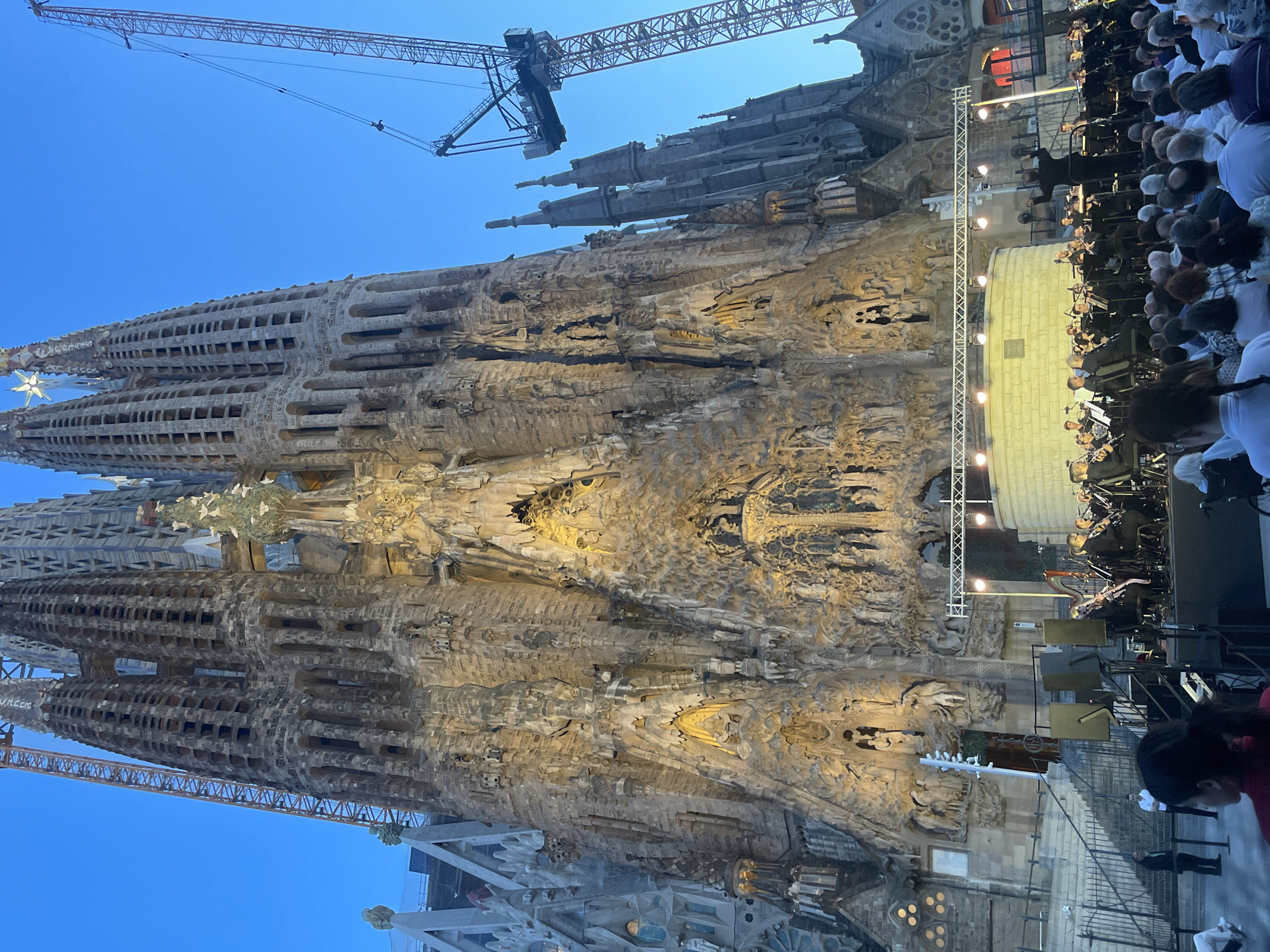 The image depicts the Sagrada Familia in Barcelona, Spain, a large, unfinished Roman Catholic minor basilica. The basilica's facade, intricately carved and illuminated, dominates the upper portion of the image. In the foreground, an orchestra is performing on a stage set up in front of the basilica. A crowd of people is seated, facing the orchestra and the basilica. Two cranes are visible in the background, indicating ongoing construction. The sky is a clear blue, suggesting it is daytime, though the lighting on the basilica indicates it might be dusk or evening. The overall scene captures an event taking place at this iconic architectural landmark.