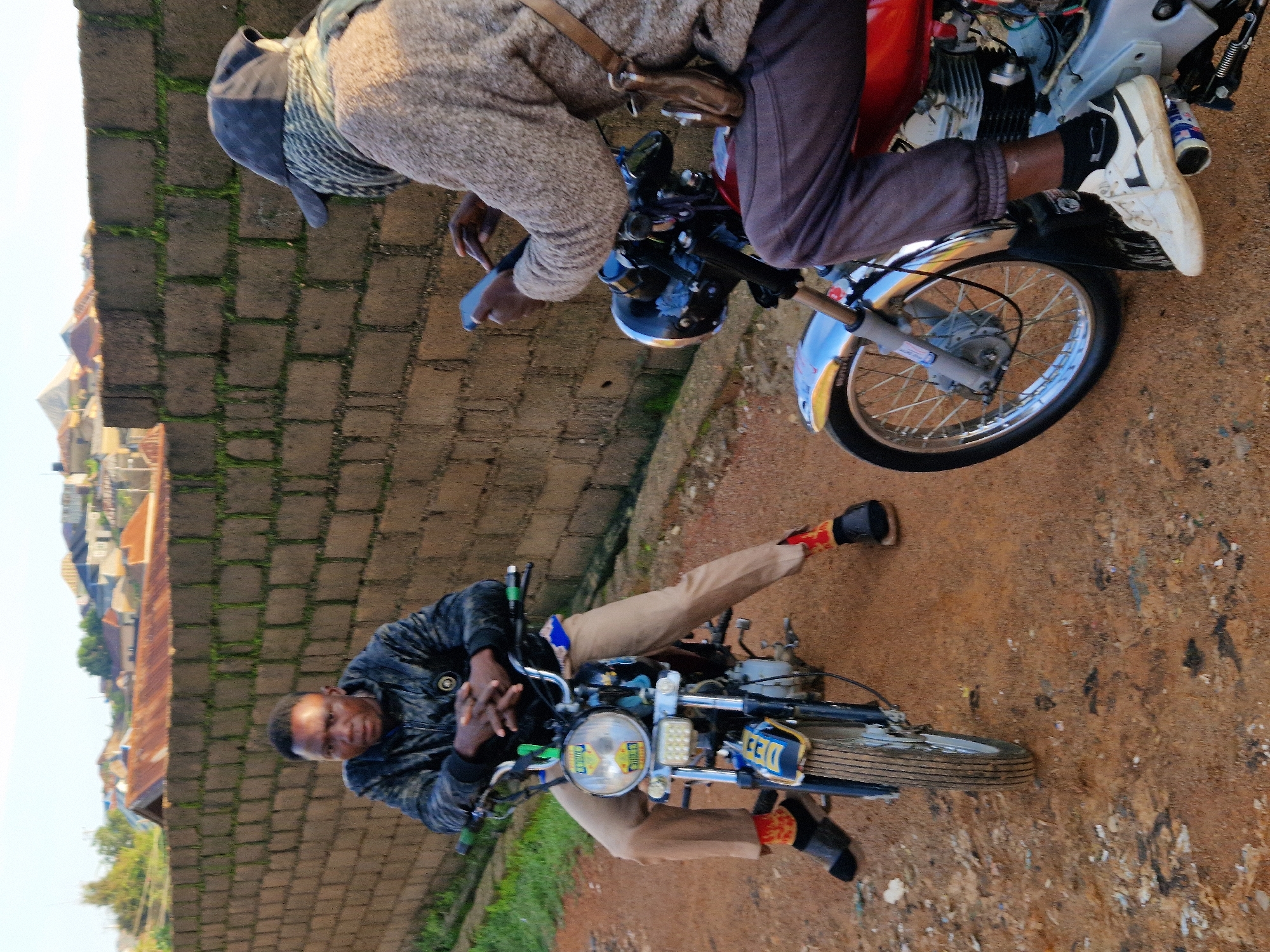 Two individuals are positioned on motorcycles in an outdoor setting with a brick wall in the background. The person on the left, facing forward, is seated on a blue and silver motorcycle, wearing a black jacket, khaki pants, and red and black socks with dark shoes. Their hands are clasped on the handlebars. The person on the right is facing away from the camera, on a red motorcycle, wearing a grey knitted sweater, dark pants, a dark cap, and a scarf. This individual is holding a blue smartphone. The ground is a dirt path. In the distance, buildings are visible on a hillside, and the sky is clear and bright.