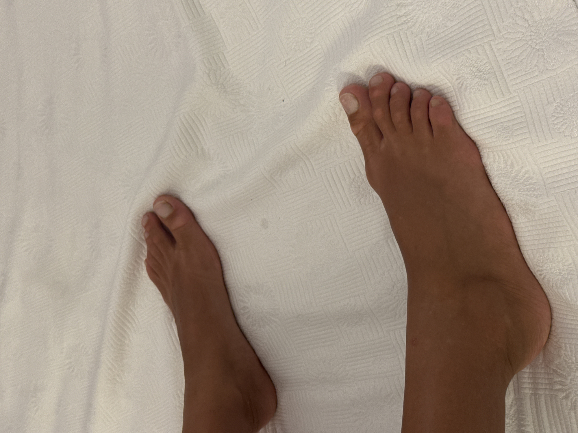 The image shows two human feet positioned on a white textured surface. The feet are tanned and appear to be bare. The surface beneath the feet has a raised geometric pattern, possibly a blanket or bedspread. The lighting suggests an indoor environment. The location of Aksu, Turkey is not visually discernible from the image.