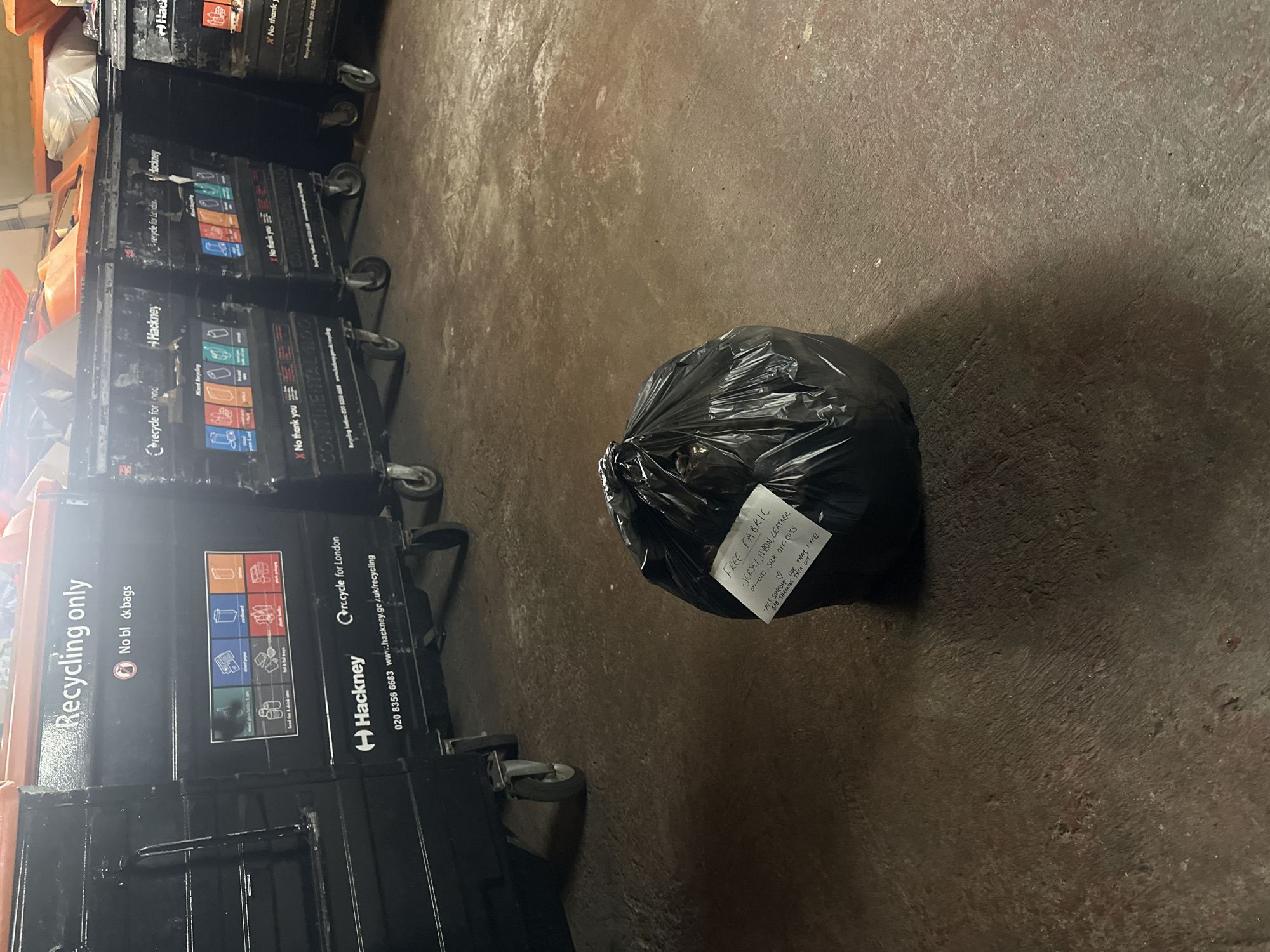 The scene shows a black plastic bag on a concrete floor, adjacent to a row of recycling bins. A handwritten note attached to the bag reads "FREE FABRIC - JERSEY, NYLON, LEATHER OFF-CUTS, SILK OFF-CUTS -PLS SOMEONE USE THEM, I FEEL BAD THROWING THEM OUT." The recycling bins have the "Hackney" logo, indicating this location is in Hackney, London, United Kingdom. The bins are labelled with recycling instructions for various materials. The bins also have a phone number and website for recycling information in Hackney.
