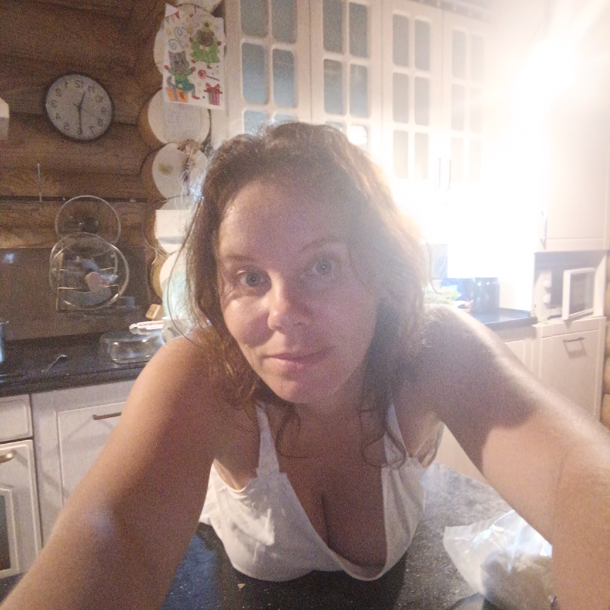 The image is a selfie taken indoors, likely in a kitchen. A woman is the primary subject, positioned in the foreground and looking directly at the camera. She has light brown hair and appears to be wearing a white top. The background shows kitchen cabinets and a microwave, suggesting a domestic setting. A clock is visible on a wooden wall in the upper left corner. The lighting is bright and appears to be coming from the right side of the frame, causing some glare. The overall scene is casual and informal. No specific location in Большой Букор, Russia, can be identified from the visual information provided.