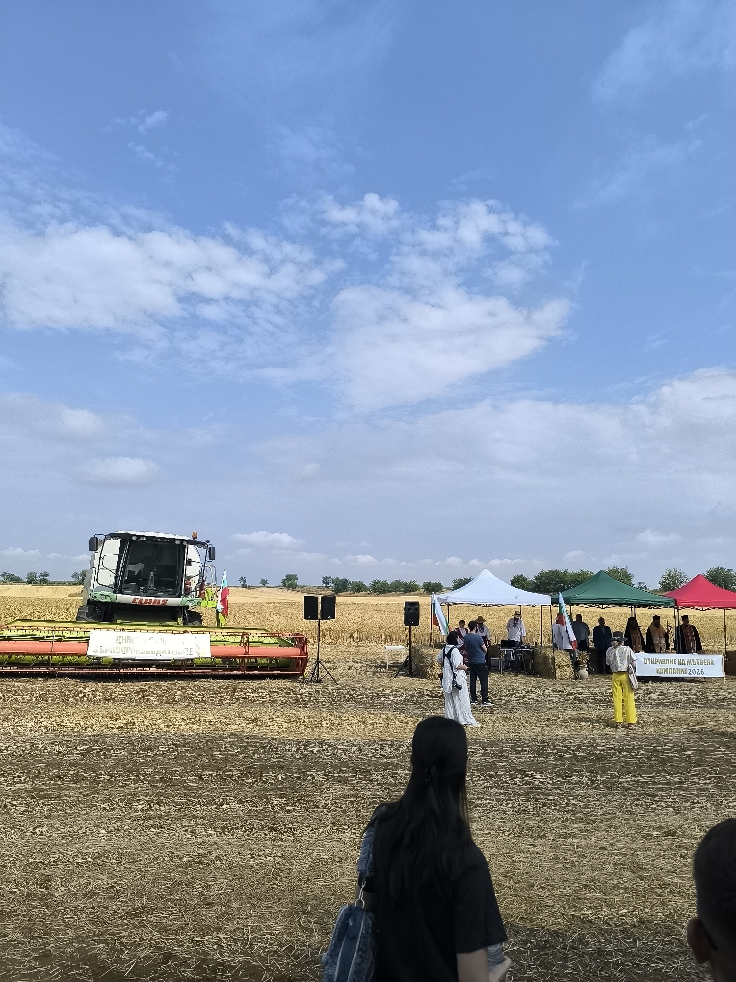 In the rural setting of Koshava, Bulgaria, a field of harvested wheat serves as the backdrop for an agricultural event. A Claas combine harvester, prominently displayed on a platform of hay bales, dominates the left side of the frame. In the middle ground, several tents – white, green, and red – provide shade for attendees and likely house exhibits or informational booths. A group of people, dressed in a mix of casual and traditional attire, are gathered near the tents. Some individuals are engaged in conversation, while others observe the proceedings. A banner with Bulgarian text, possibly announcing the event or a specific campaign, is visible on the right side of the tents. The sky above is a clear blue, dotted with scattered white clouds, suggesting fair weather. The ground is covered with dry wheat stubble, indicating the harvest has recently concluded.