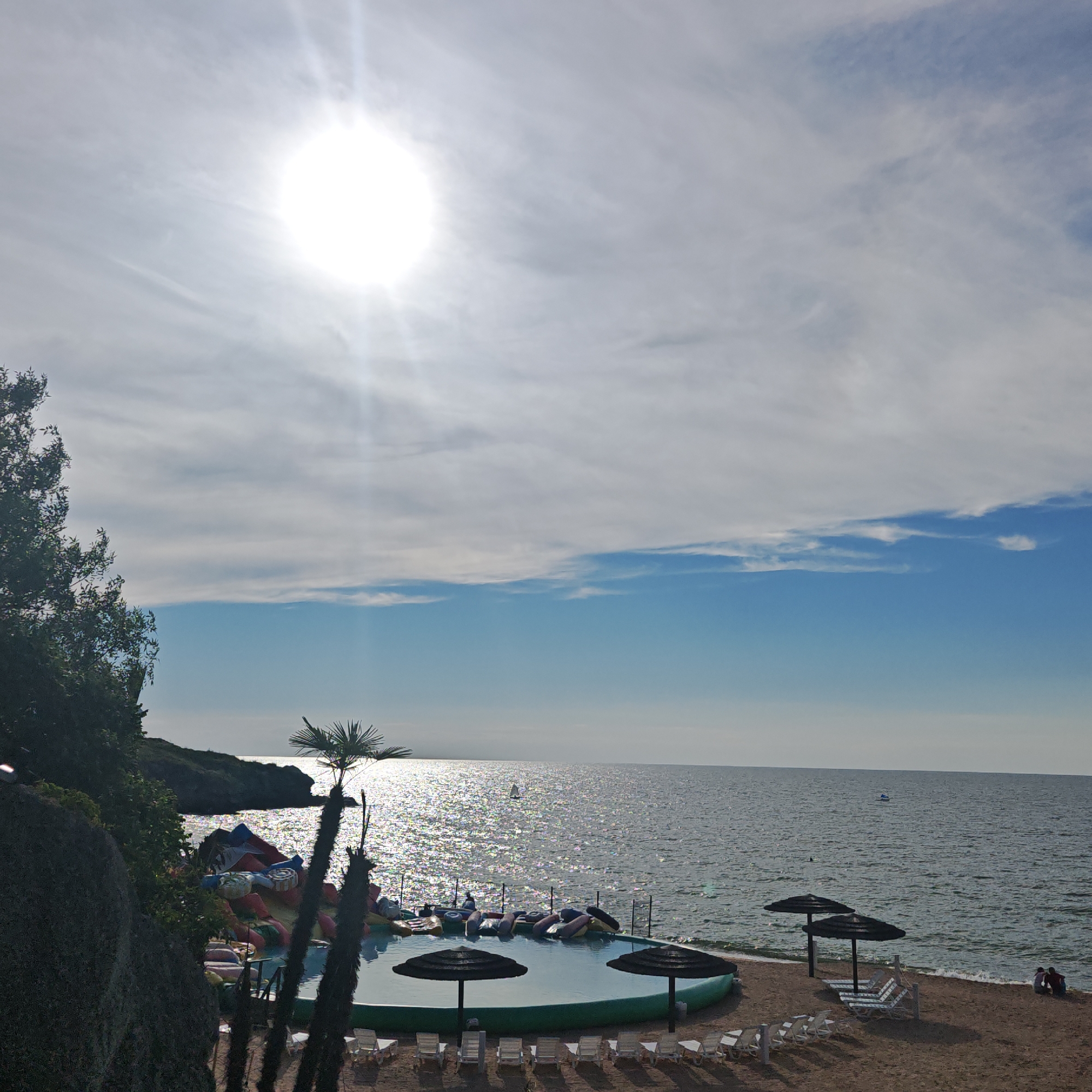 The image depicts a sunny day at a beachfront recreational area in Мысовское сельское поселение, Ukraine. The sky is bright with the sun high and prominent, casting a strong glare on the water. Wispy clouds are scattered across the blue sky. In the foreground, a rocky incline covered in dense green foliage is visible on the left. A swimming pool with inflatable toys and a water slide is situated on the sand, with several lounge chairs and umbrellas around it. The sea stretches out to the horizon, with a few small boats visible. Two people are seated on the sand by the water's edge in the lower right corner of the image. The overall scene suggests a resort or public beach area designed for leisure and recreation.
