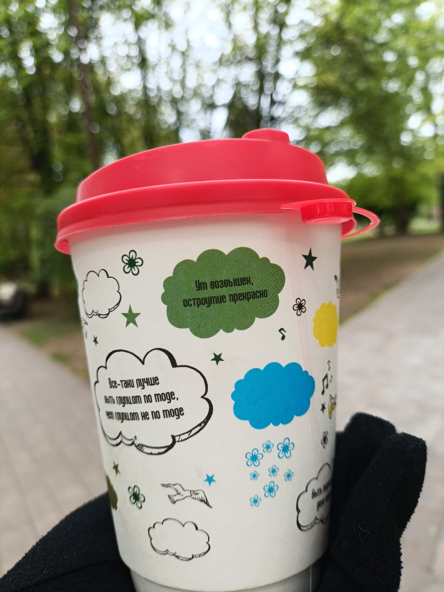A person holds a white paper cup with a red lid, adorned with colorful cartoonish designs. The cup has cloud-shaped text bubbles, one of which contains the phrase "Все-таки лучше быть глупцот по моде, чет глупцот не по моде" (which translates to "It's still better to be stupid in fashion than to be stupid out of fashion"), and another saying "Ут возвышен, остроумие прекрасно" (which translates to "Elevated mind, wit is wonderful"). The background is blurred, suggesting an outdoor location, likely a park or a sidewalk, with trees visible in the distance. The setting appears to be in the city of Kaliningrad, Russia. The cup is held by a hand wearing a black glove.