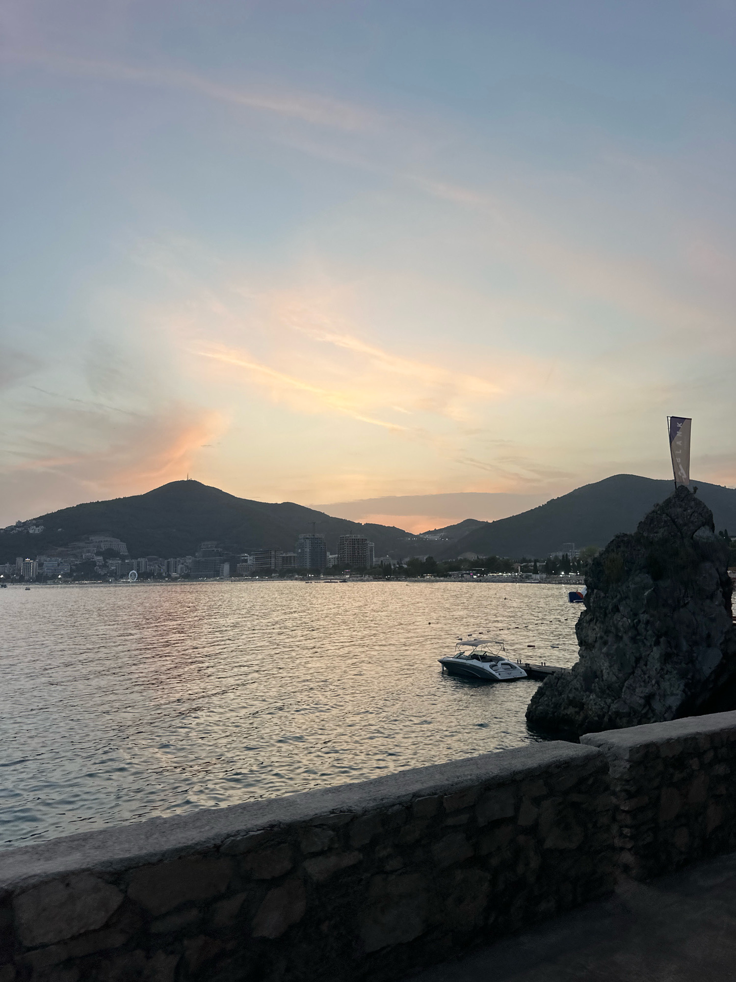 The image depicts a serene sunset scene over the Adriatic Sea in Budva, Montenegro. In the foreground, a stone wall lines a walkway, with rough-hewn stones forming the base and a smooth concrete top. Beyond the wall, the calm sea reflects the soft hues of the sky. To the right, a cluster of large, dark rocks rises from the water, topped with a flag displaying a logo. A small, white motorboat is moored to a dock near the rocks. In the mid-ground, the coastline of Budva stretches across the horizon, featuring a collection of buildings, including hotels and residential structures, nestled against a backdrop of verdant, rolling hills. A prominent hill with a telecommunication tower on its summit dominates the left side of the cityscape. The sky above is painted with soft pastel shades of pink, orange, and blue, with thin, wispy clouds suggesting the end of daylight. The overall atmosphere is peaceful and picturesque, capturing the beauty of the Montenegrin coast at dusk.