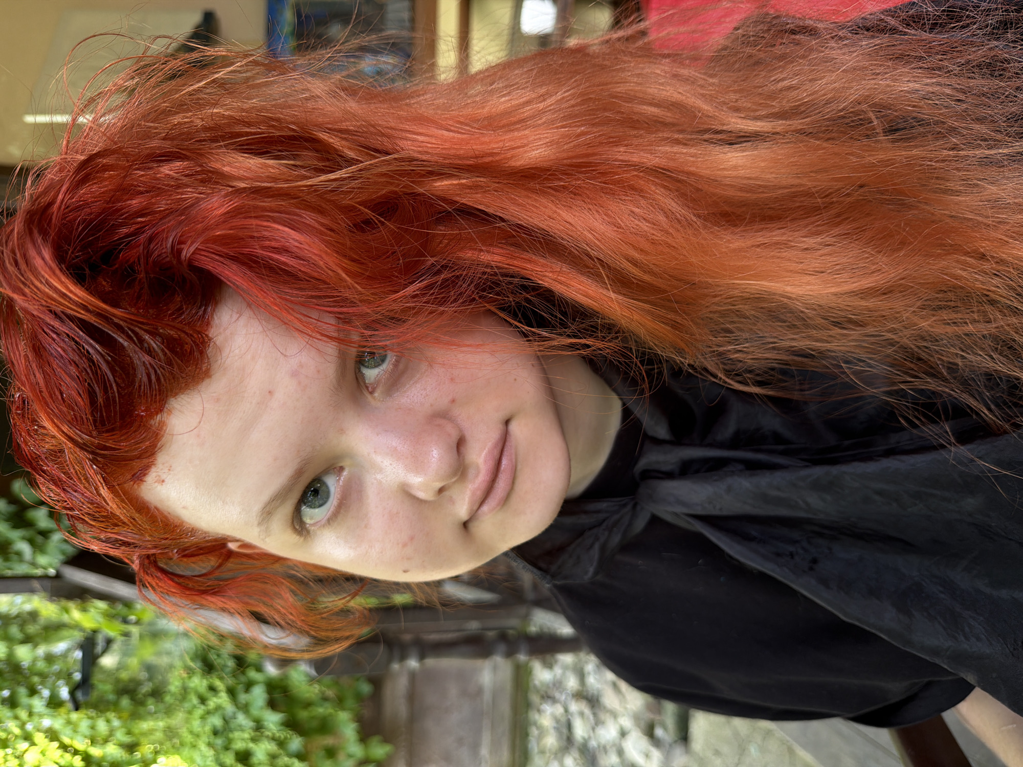 A close-up, waist-up outdoor portrait shows a young person with bright red, wavy hair that is partially covering their face. They are wearing a black garment that appears to be a cape or smock, tied at the neck with a black ribbon. The person has fair skin with some freckles and is looking towards the camera with a neutral expression. The background is blurred, suggesting an outdoor setting with greenery and possibly some wooden structures, consistent with a garden or patio area. The lighting indicates it is daytime. The location in Sochi, Russia, is not visually identifiable from the image alone; this detail is external information.
