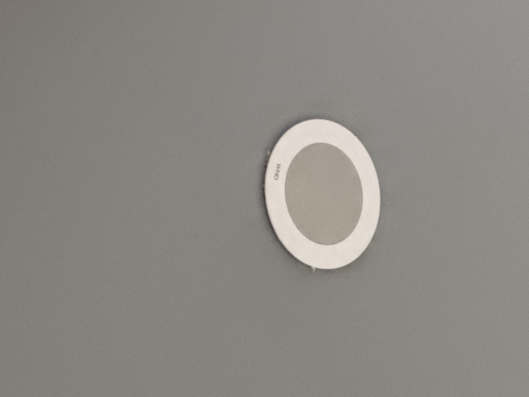 The image shows a plain, uniformly grey wall with a slight textured finish. Mounted on this wall is a circular device, positioned slightly to the right of the center of the frame. The device consists of a white outer ring and a light grey inner circle. On the white outer ring, the letters "WHO" are visible, printed in black and oriented vertically. There are no people, activities, or distinct location cues beyond the wall and the device itself. The lighting appears even, suggesting an indoor setting. The scene is static, devoid of any discernible event or emotion. The image depicts a detail from Yerevan, Armenia.