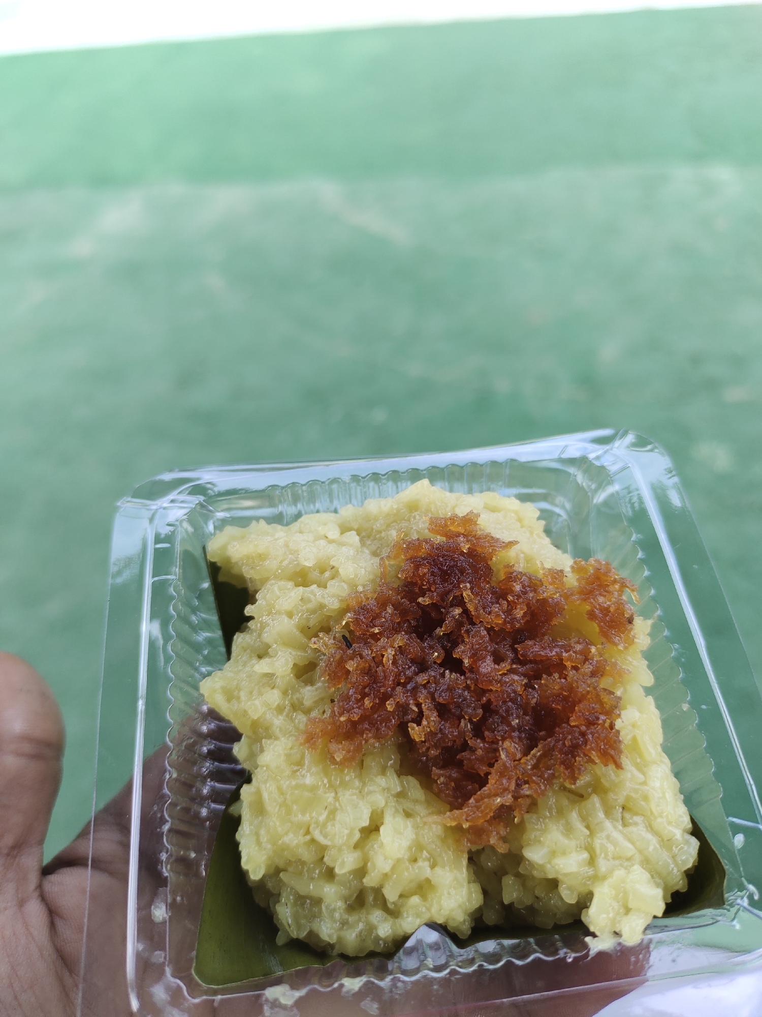 A human left hand holds a clear plastic container containing yellow sticky rice topped with dark brown, shredded material, located in Meunasahaluedrienlhoksukon Barat, Indonesia. The rice, molded into a rectangular shape, rests on a segment of green banana leaf within the container. The brown topping exhibits a fibrous texture, consistent with sweetened coconut floss. The transparent plastic container features vertical ribbing along its lower sides. The background is a heavily blurred, uniform green surface.
