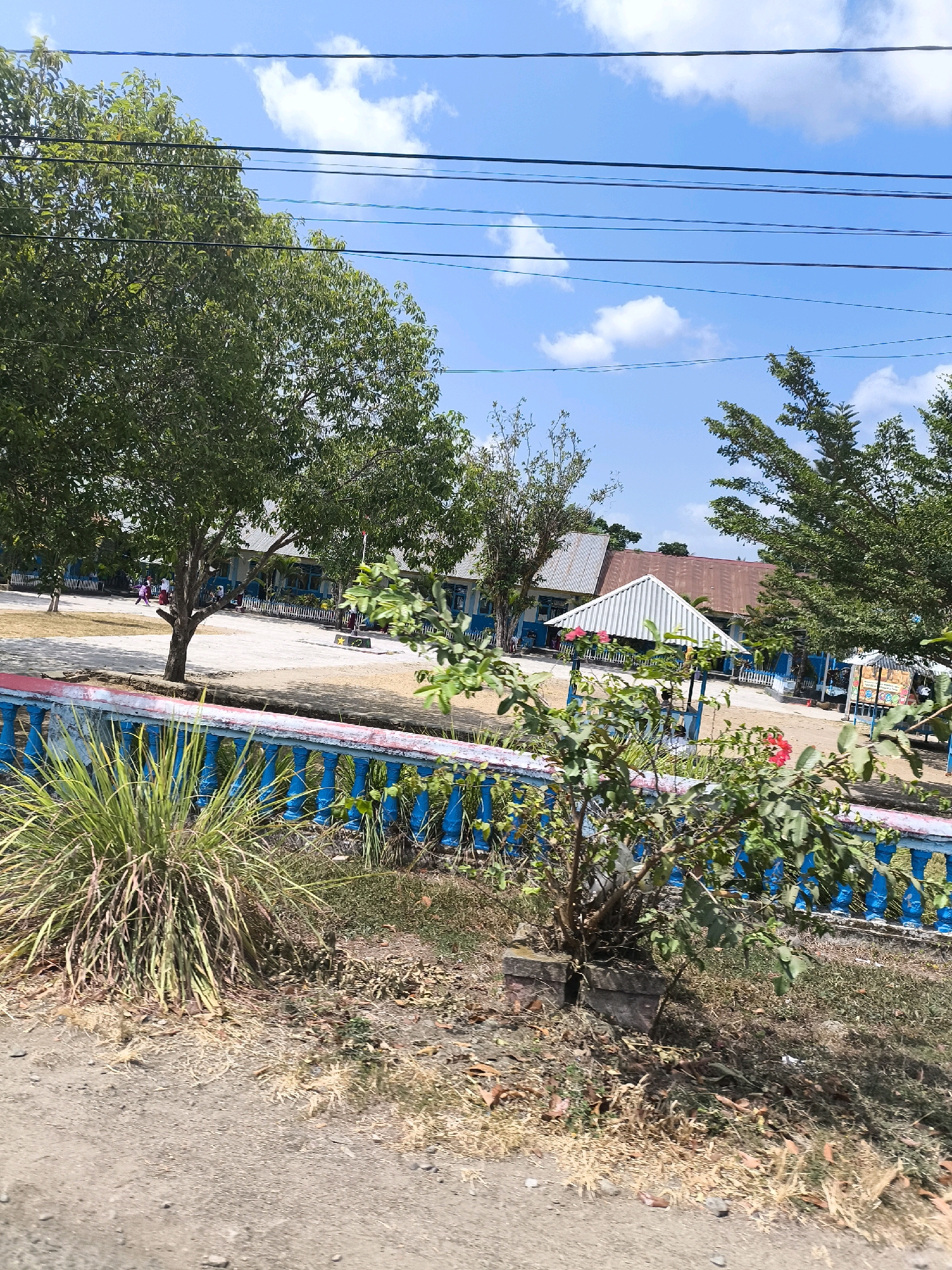 This image depicts a scene in the city of Sido Mulyo, Indonesia. In the foreground, a dirt path runs along the bottom of the frame, bordered by dry grasses and fallen leaves. A bright blue fence with red trim runs horizontally across the middle ground, separating the path from a schoolyard. Beyond the fence, a school building with blue walls and light-colored roofs is visible. Several trees with green foliage are scattered throughout the schoolyard. Children in uniforms can be seen walking and standing in the schoolyard. Overhead, power lines stretch across a bright blue sky with scattered white clouds.