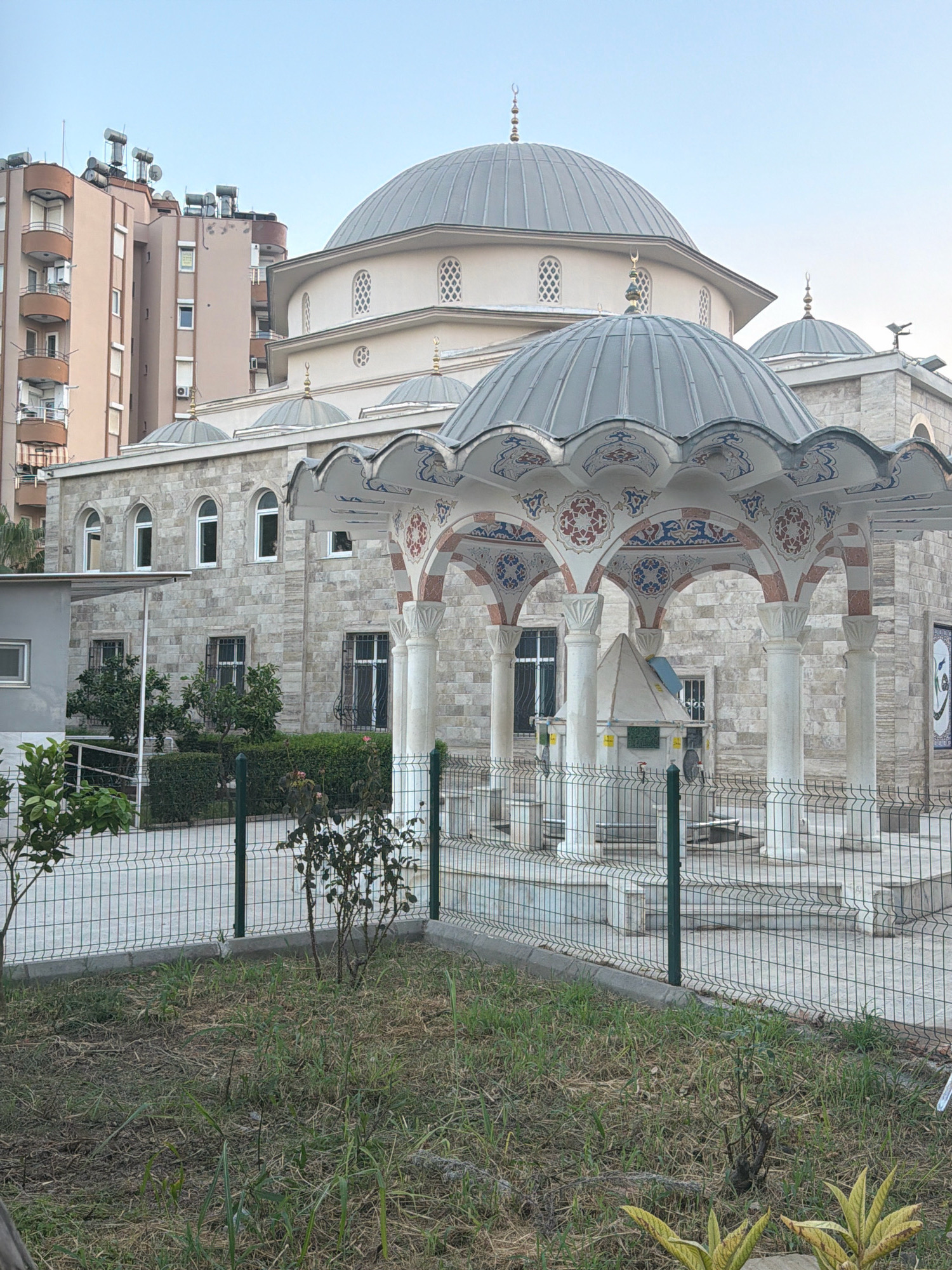 An exterior, ground-level, eye-level shot depicts a mosque with a large dome and several smaller domes, constructed with light-colored stone blocks. The mosque features arched windows and is surrounded by a fence. In front of the mosque is a covered area with decorative arches supported by columns. A small, pyramidal structure with a faucet is visible within this area, suggesting it might be a fountain or ablution area. To the left of the mosque, a modern, multi-story apartment building with balconies and solar water heaters on its roof stands prominently. The foreground shows a patch of dry grass and a small tree, suggesting an urban green space. The sky is clear and blue, indicating daytime. The scene appears to be located in Antalya, Turkey, based on the architectural style and building types. No people are visible in the image.