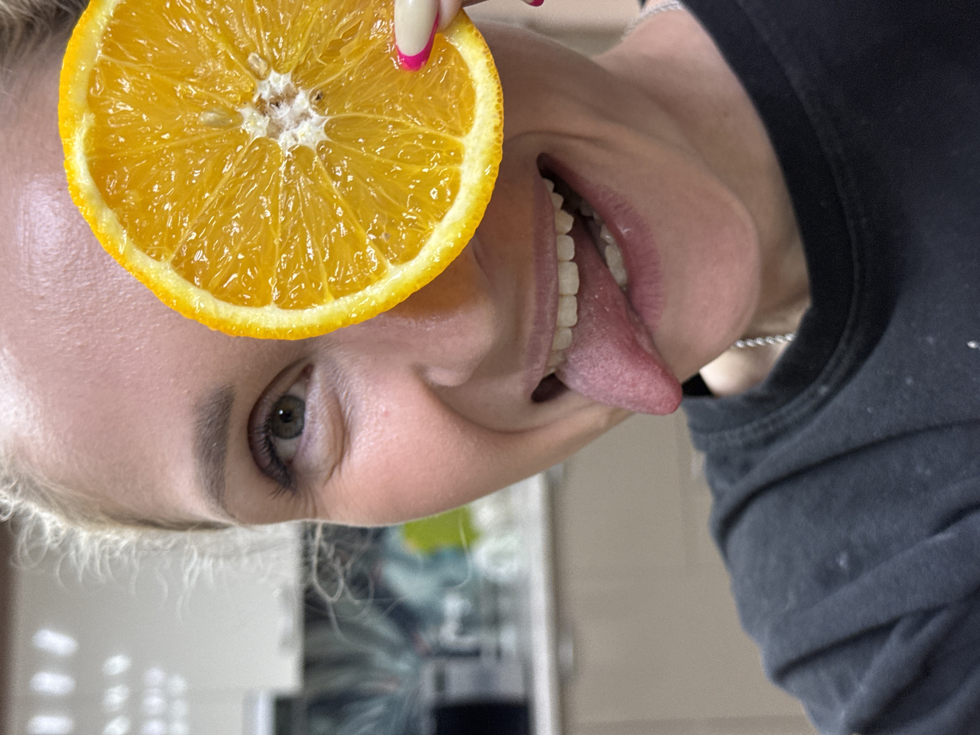 A fair-skinned person with light hair holds a halved orange over their right eye. The person's tongue protrudes from their mouth. They are wearing a dark-colored shirt and a delicate silver chain. The environment appears to be indoors, with a blurred background suggesting a kitchen or similar domestic space. The location of Chaikovsky, Russia, is not discernible from the visual information in this image.