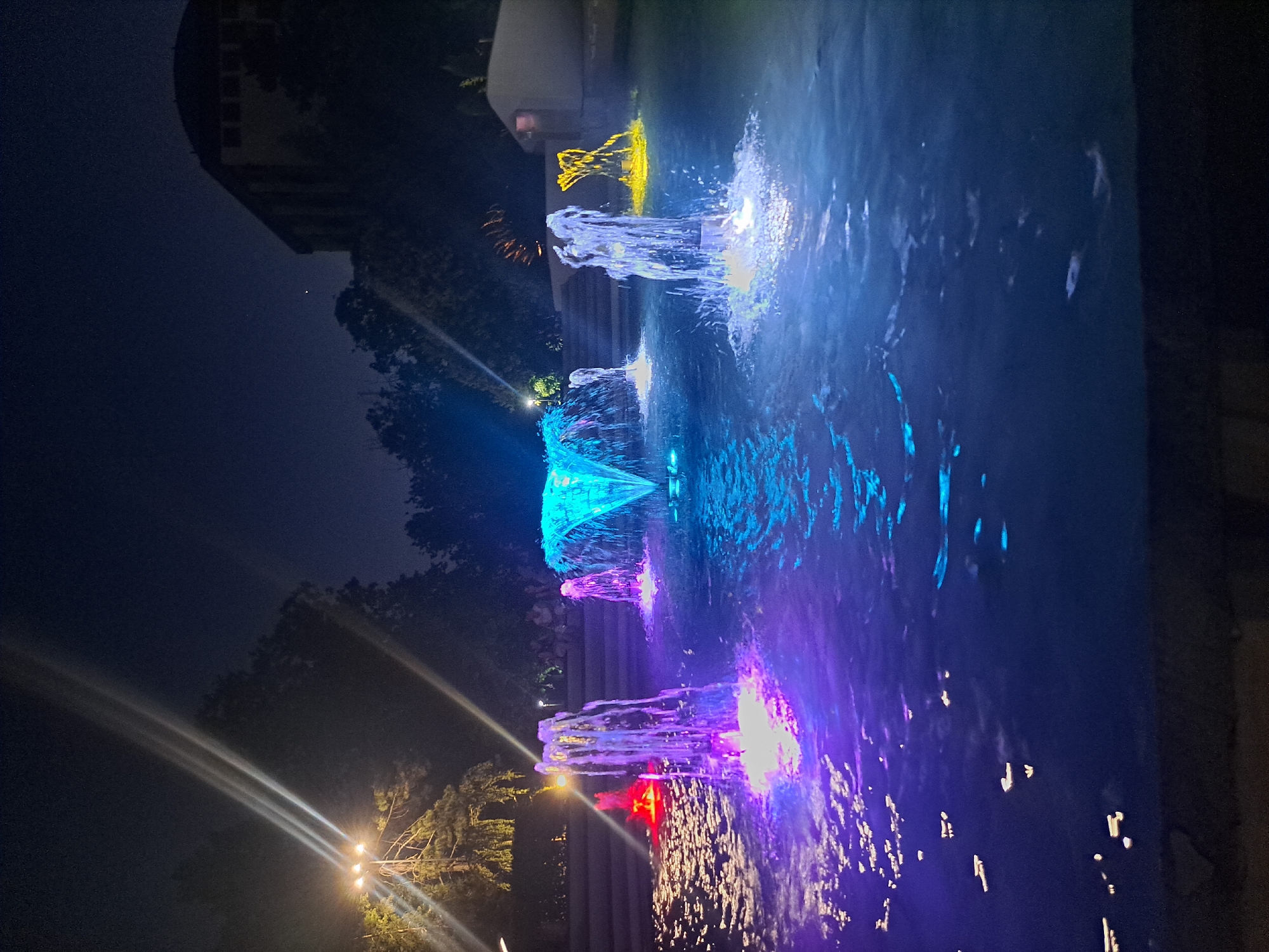 An exterior, nighttime, eye-level shot captures a brightly lit fountain display in a public area in the city of Popovo, Bulgaria. Several fountains are active, with water jets illuminated in vibrant colors of blue, purple, and yellow. In the background, trees and a building are visible under the dark night sky. The scene appears to be a park or plaza, with a few indistinct figures seated in the background. The water of the fountain is rippling and reflecting the colorful lights, creating an energetic and lively atmosphere.