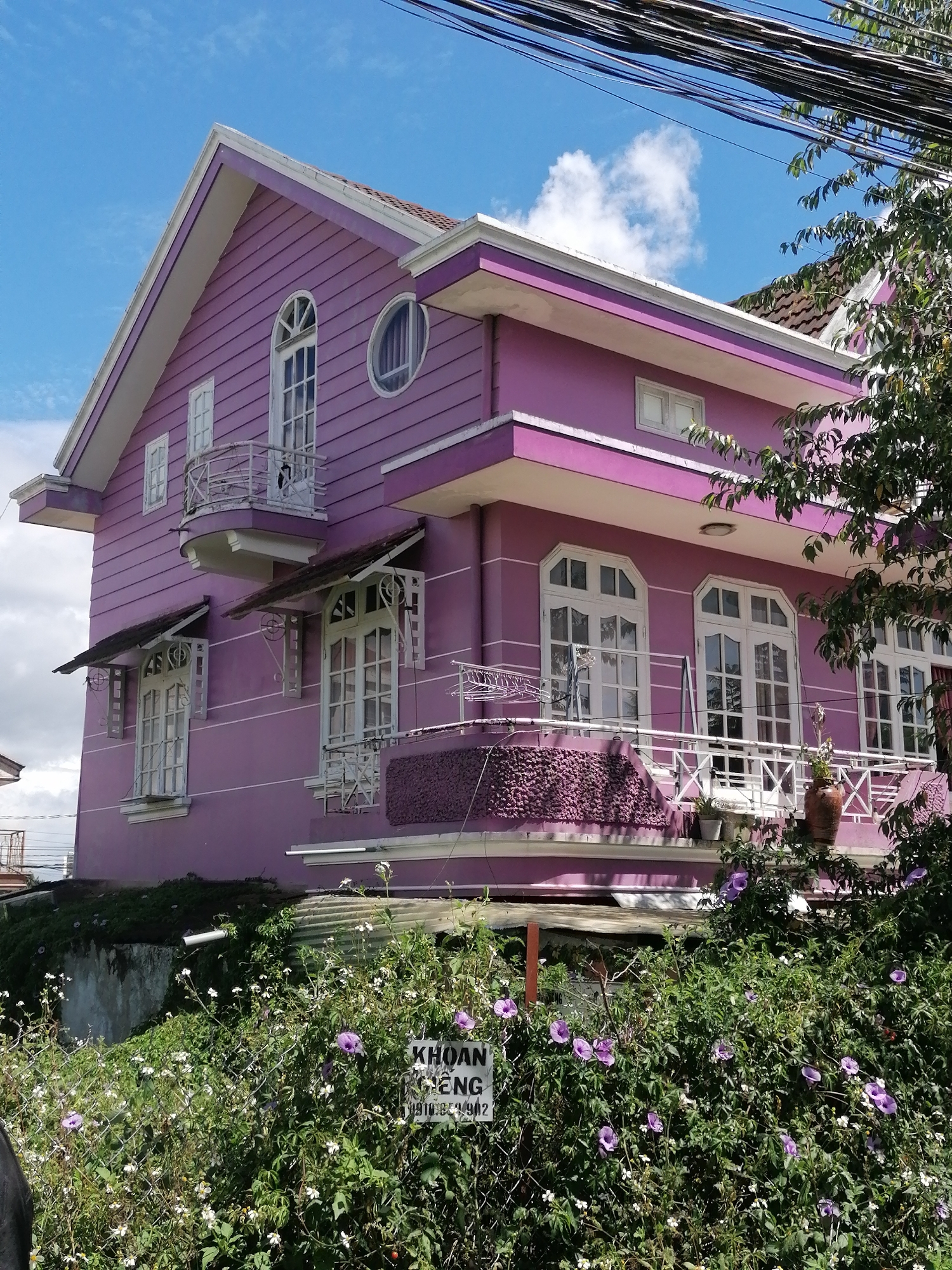 The image shows a two-story purple house with white trim and decorative balconies, located in Lâm Viên - Đà Lạt, Vietnam. The house has several large windows with white frames and intricate detailing. Lush green foliage and blooming purple and white flowers surround the base of the house, with some of the plants climbing over a chain-link fence. A sign with Vietnamese text is visible among the greenery. The sky is blue with scattered white clouds, and overhead, electrical wires are present. The scene suggests a residential setting with a vibrant, somewhat whimsical architectural style.