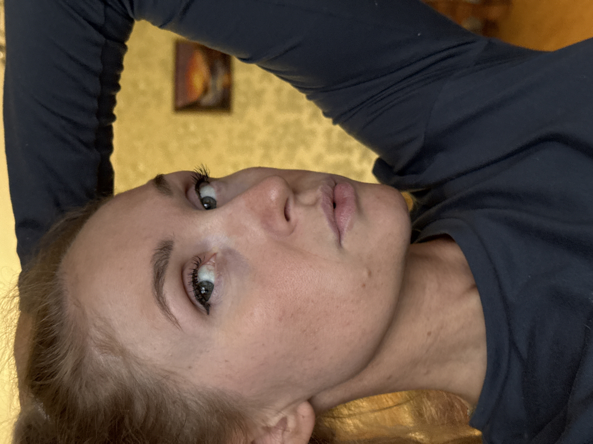 A woman with fair skin and light brown hair is positioned at an angle in the frame, looking upwards. She is wearing a dark blue long-sleeved shirt. Her left arm is raised behind her head. Her eyes are blue-grey, and she has light-colored eyelashes. The background appears to be a gold-toned wall with a framed painting. It is not possible to determine the location as Perm, Russia from the image alone.