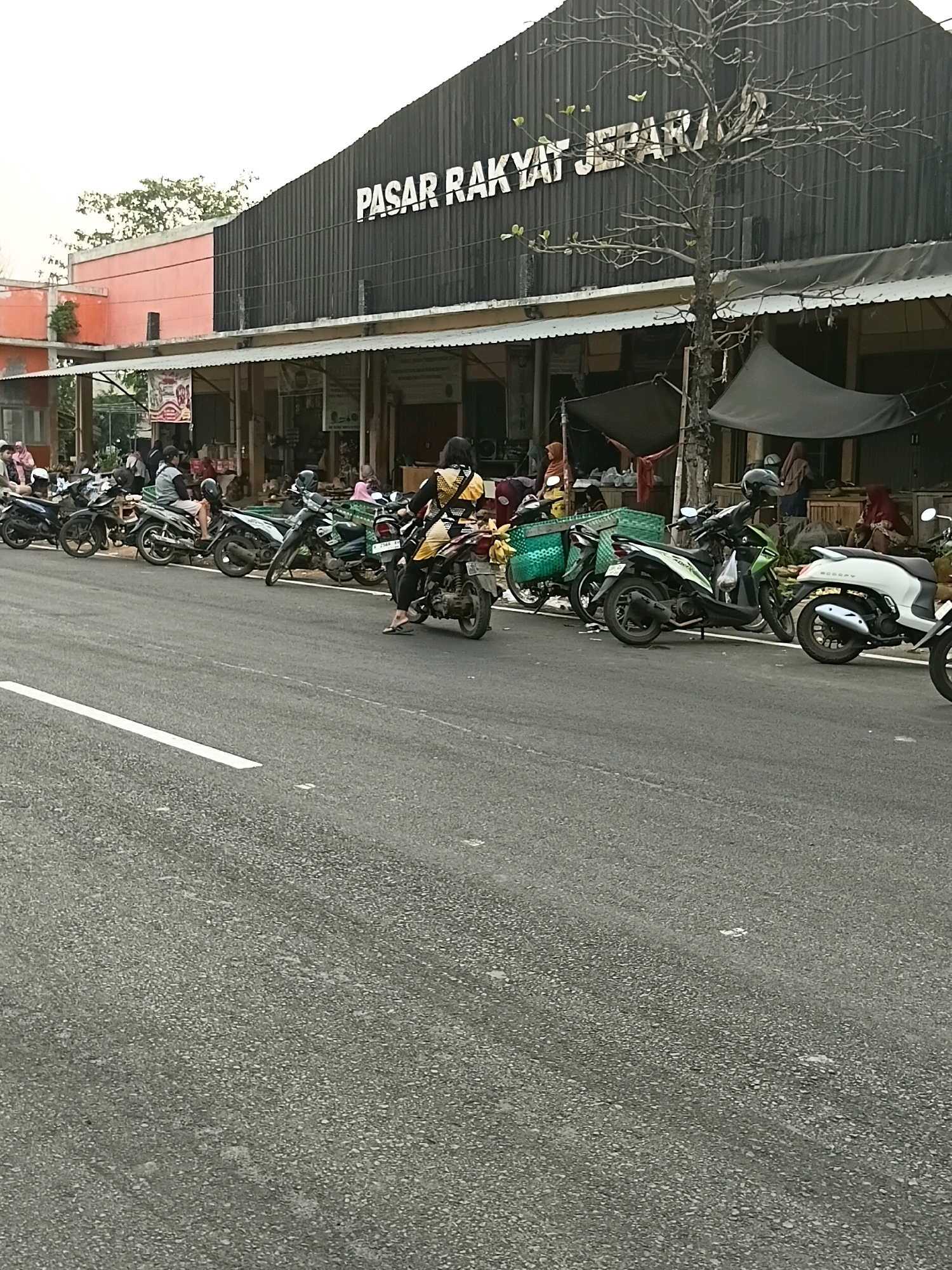 The image depicts a street scene in front of a building with the sign "PASAR RAKYAT JEPARA" indicating it is a public market in Jepara, Indonesia. A row of parked motorcycles is visible along the side of the road. Several individuals are present, some on or near the motorcycles, and others in the background near market stalls. The market appears to have awnings or temporary structures set up. The road is paved and has a white lane marking. The overall lighting suggests it is daytime.