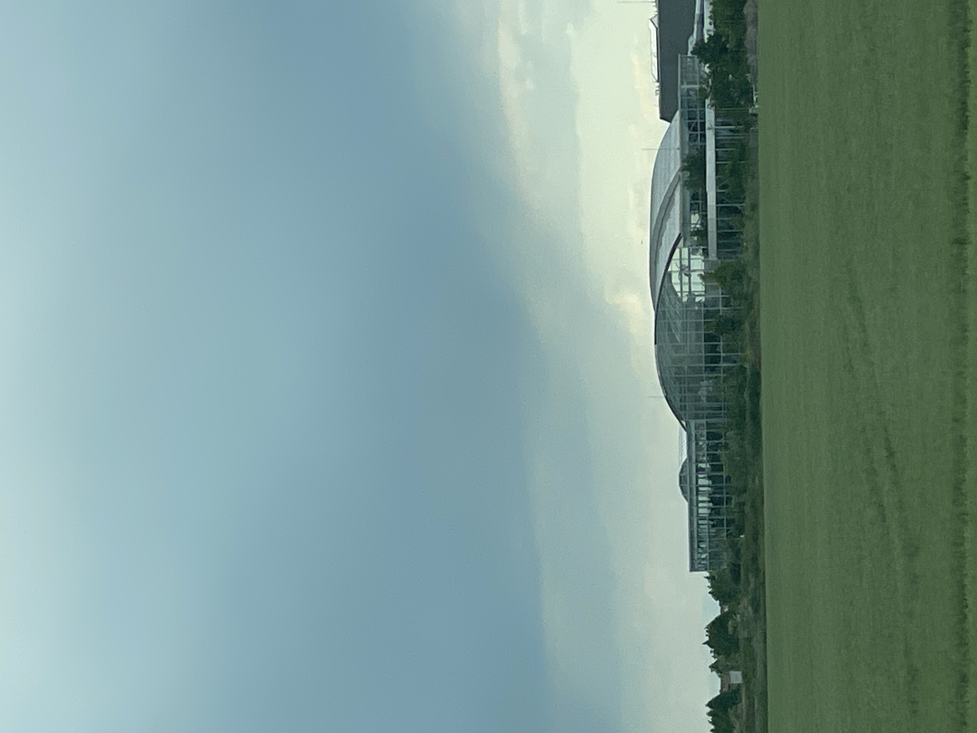 The image depicts a landscape in Balotești, Romania. The left side features a light blue sky with wispy clouds. Adjacent to the sky, in the middle ground, is a modern architectural structure resembling a botanical garden or greenhouse. This structure is characterized by its curved glass facade and grid-like framework, and contains vegetation. To the right of the structure lies a flat, green field of grass. In the background, there are hints of additional buildings and greenery.