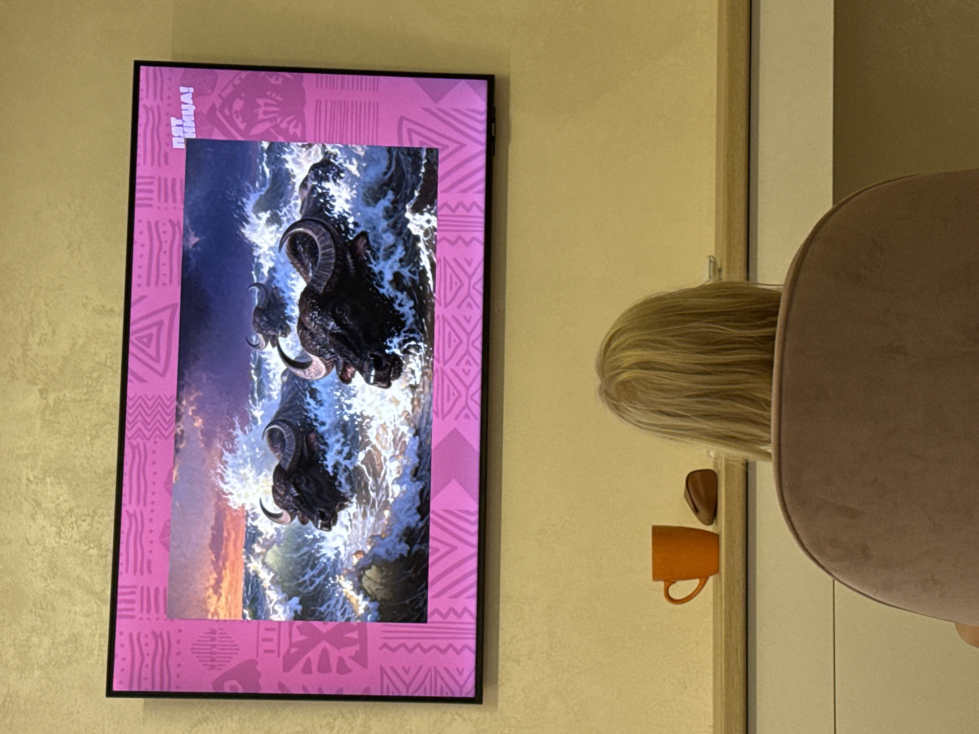 A horizontal, medium shot captures a television screen displaying an image of water buffaloes swimming in rough seas, set against a backdrop of a vibrant sunset. The television is mounted on a textured, yellow wall. The screen's border features a pink, patterned frame. To the left of the television, on a shelf, sits an orange mug and a small, brown object, possibly a piece of bread. In the foreground, the back of a person with blonde hair is visible, seated in a plush, pinkish-brown chair, facing the television. The scene appears to be an indoor setting, likely a living room, with the television being the central focus. The image's composition suggests a casual viewing experience, possibly in a residential apartment in Sevastopol, Ukraine.