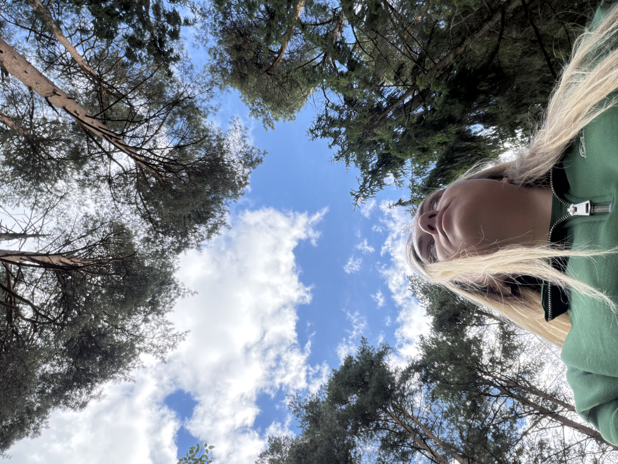 The image is a low-angle shot taken from the ground, looking up towards a sky with scattered white clouds and patches of blue. Tree branches and foliage surround the frame, creating a circular border around the sky. A fair-skinned person is positioned on the right side of the frame, partially obscuring the view, wearing a green zip-up jacket. Their face is visible, with eyes looking upward. The location may be in a wooded area or park in Votkinsk, Russia, based on the presence of trees and sky.