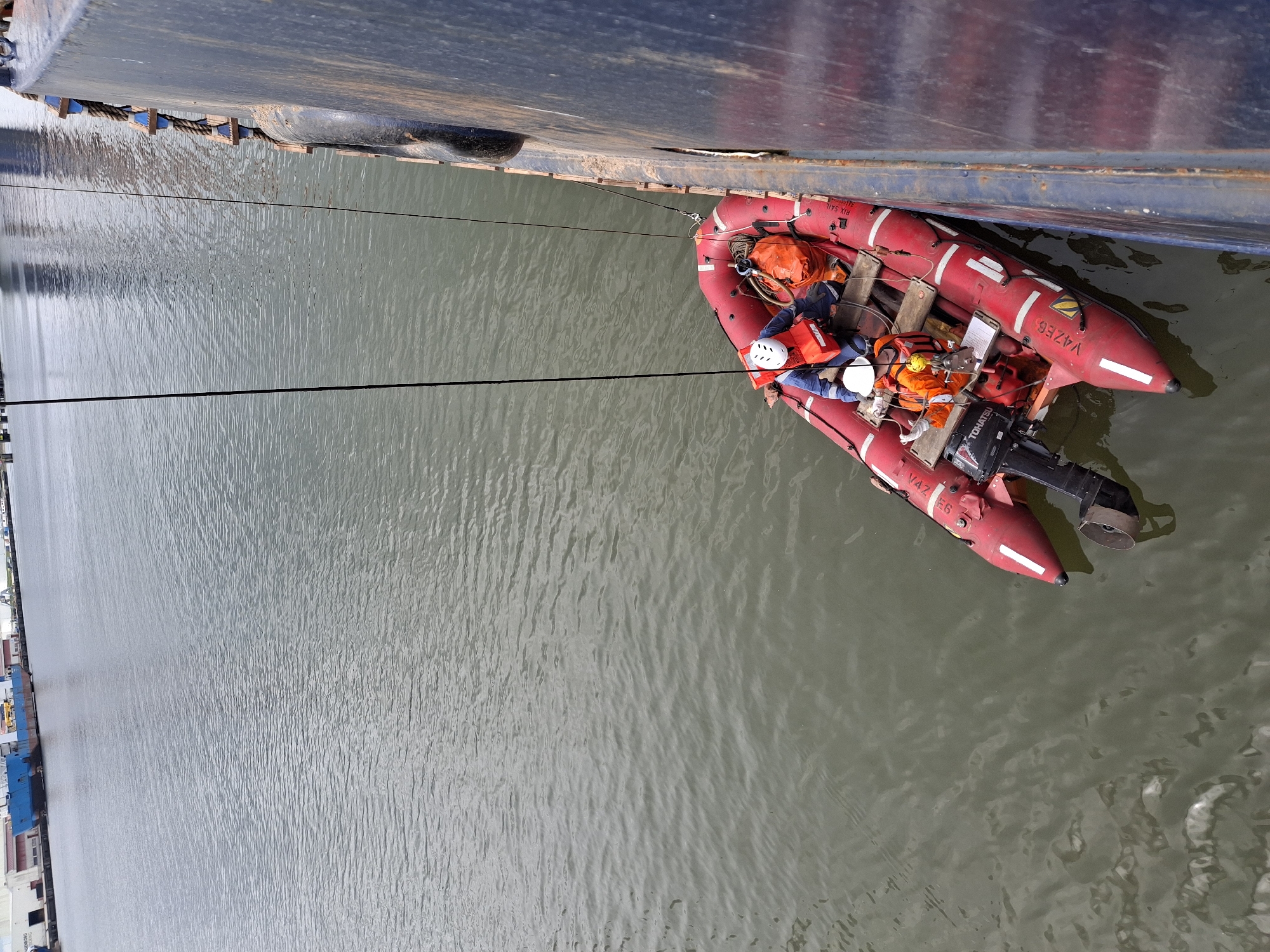 A red inflatable boat, carrying two individuals, is alongside a large ship or vessel. The individuals in the boat are wearing helmets and life vests. The boat is secured to the larger vessel by ropes. The environment is a body of water, possibly a river or harbor, with reflective surface. Along the river are rows of container warehouses.