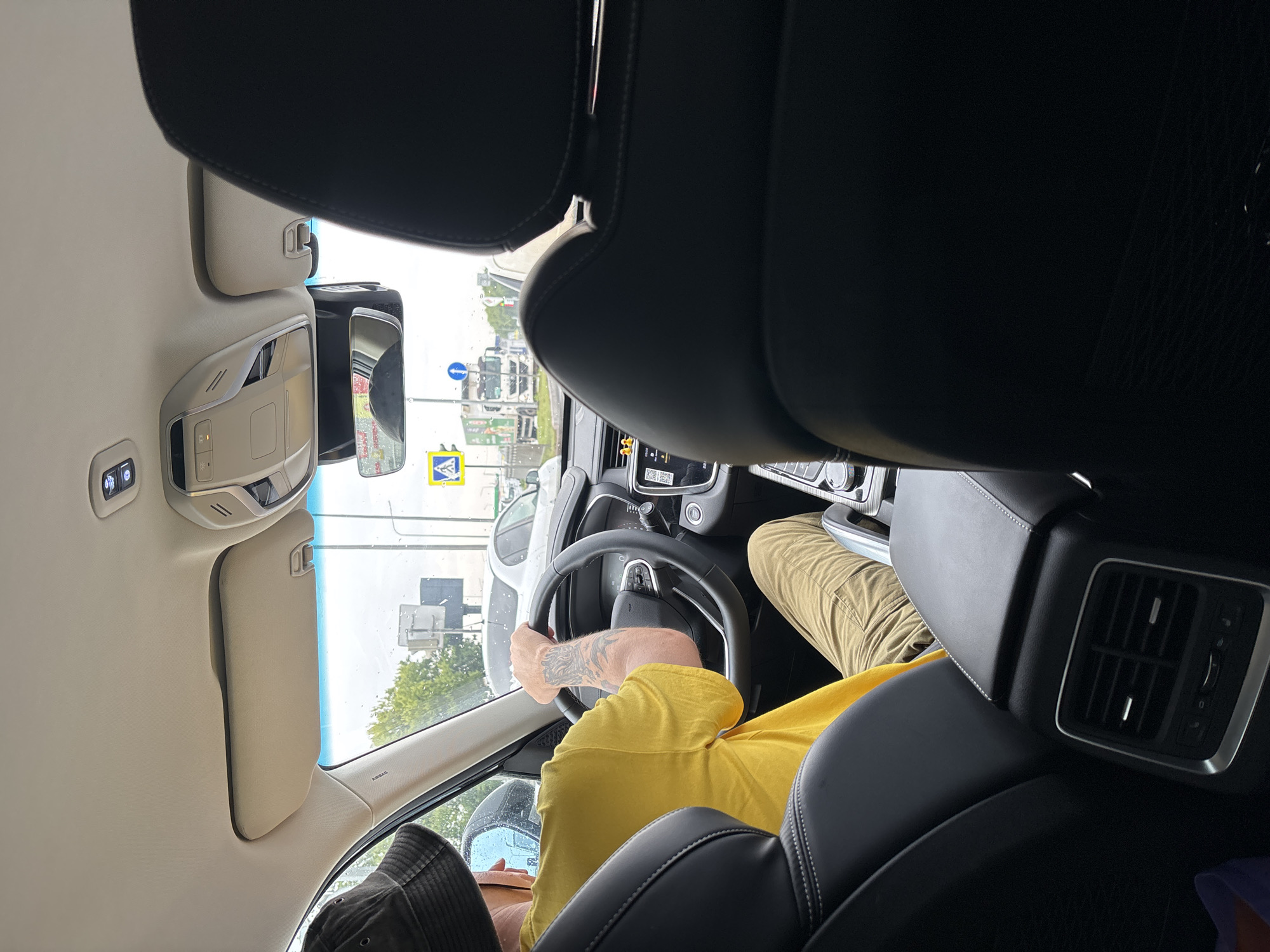 The image shows the interior of a car, viewed from the back seat, looking forward. A person wearing a yellow t-shirt and a bucket hat is driving the car. They have tattoos on their right arm. The car is on a city street, with other vehicles and traffic signals visible outside the windows. The sky is overcast, and it appears to be daytime. There are buildings and trees in the background. The car's dashboard and steering wheel are visible. A rearview mirror is positioned above the dashboard. The overall scene suggests a person driving in a built-up area, possibly during a commute or a trip. The presence of traffic signs and other vehicles indicates an urban environment. The weather appears to be cloudy or rainy.