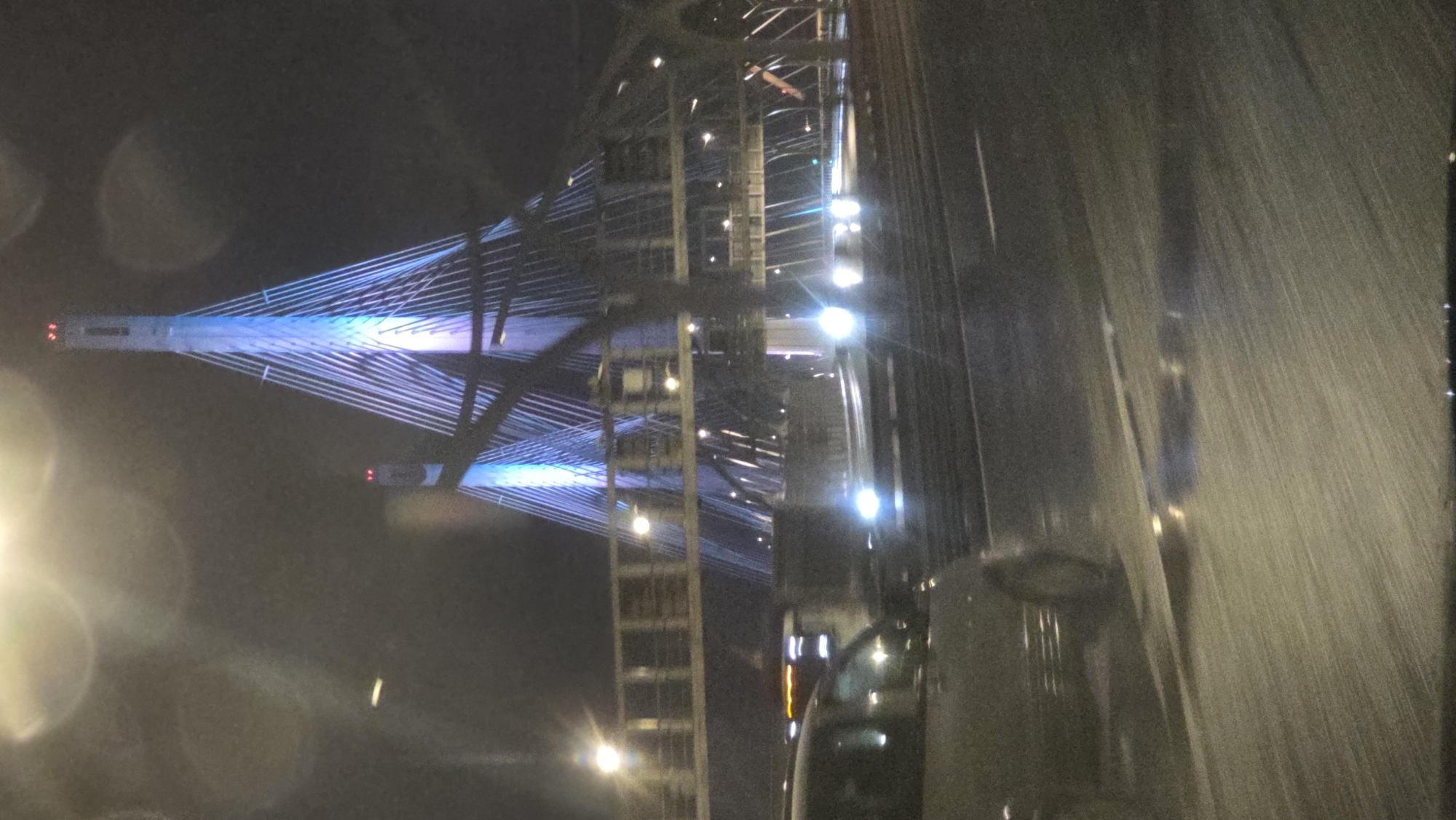 A nighttime scene depicts a portion of the Saint Petersburg, Russia cable-stayed bridge illuminated with blue lights. The image appears to be taken from inside a vehicle, evidenced by the water streaks on the glass. A section of the bridge's support structure with lights is visible on the right of the image.