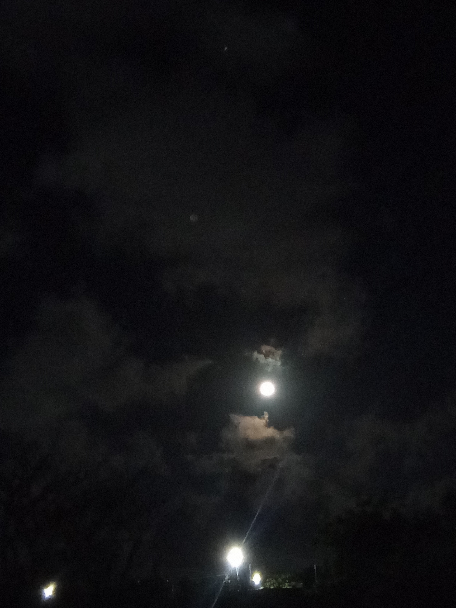 The image shows a nighttime scene with a bright moon partially obscured by clouds. The sky is dark, with visible cloud cover and a couple of fainter, unidentified light points. In the lower portion of the image, there are two bright streetlights illuminating foliage. It's impossible to verify the location with absolute certainty using just the visual information.
