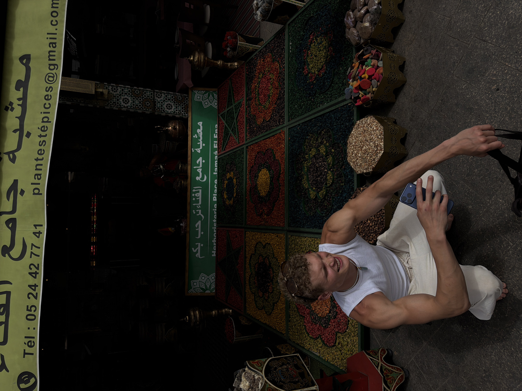 The image depicts a young man crouched on the ground in a market in Marrakesh, Morocco, likely in or near the Jemaa el-Fna square. He is wearing a white tank top and loose-fitting cream-colored pants, with sunglasses pushed up on his curly hair. He is holding a blue smartphone in his right hand and a black bag in his left.
In the background, there is a vibrant display of dried spices or herbs arranged in decorative patterns on a large mat. The mat is divided into square sections, each featuring a different colorful design. Above the display, a green banner with Arabic and French text reads "Herboristerie Place Jemaa El Fna," indicating the establishment's name and location. Various containers filled with different colored items, possibly spices or crafts, are visible around the man and the display. The overall scene suggests a bustling marketplace environment with a focus on traditional Moroccan goods. The lighting is dim, with some areas illuminated and others in shadow.