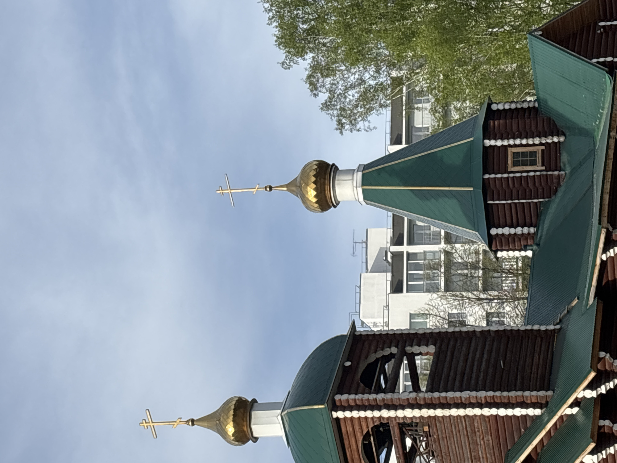 The image shows a section of a wooden church with green roofs and golden domes, each topped with a cross. The structure is detailed with decorative trim and small windows. In the background, a modern white building rises behind the church, with visible windows and architectural features. A tree with green leaves partially obscures the top portion of the building. The sky is blue with some clouds. This scene depicts the Church of George the Victorious in Chaikovsky, Russia.