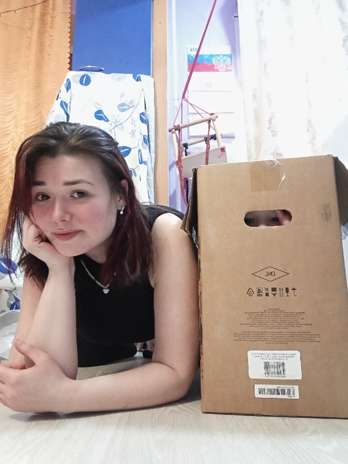 A woman rests on the floor, head propped on her hand, with a cardboard box beside her. Two eyes are visible through a hole cut in the box. The woman has dark hair, fair skin, and wears a black top. The box is brown with printed symbols and a barcode sticker. A child's swing hangs in the background against a white curtain. A patterned cloth drapes over something behind the woman.