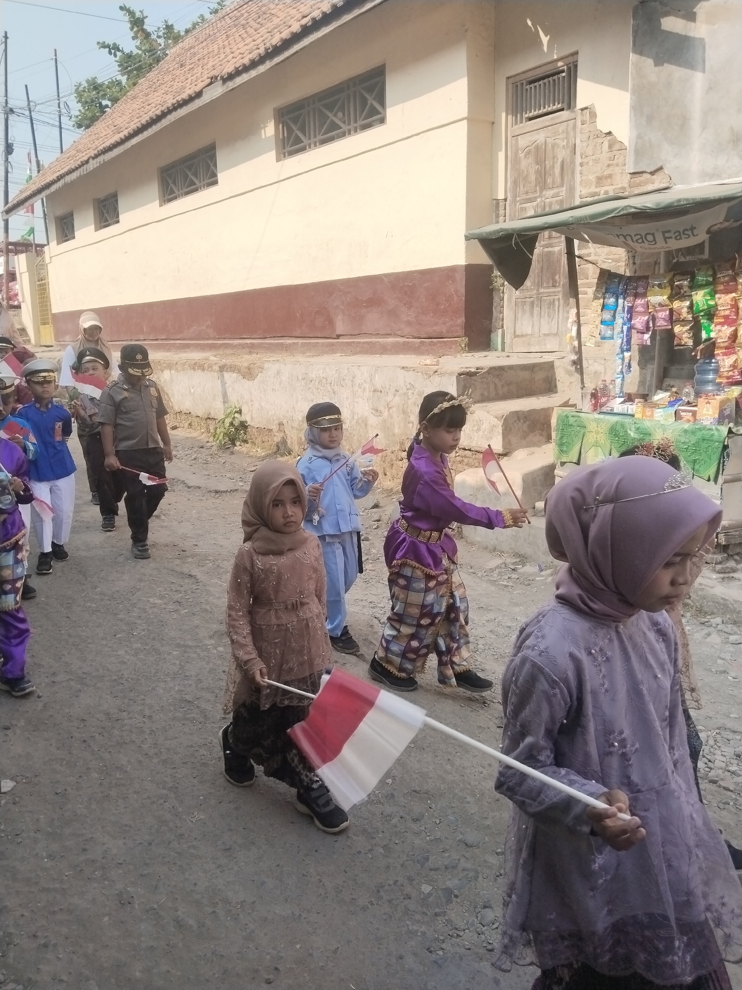 In Jembayat, Indonesia, a group of children are participating in a parade, holding Indonesian flags. The parade is taking place on a dirt road in front of a building with a terracotta tiled roof and light-colored walls. Some of the children are dressed in traditional attire, including purple outfits with intricate patterns, while others wear uniforms. One child in the foreground is holding a red and white Indonesian flag. In the background, there is a small shop with colorful merchandise displayed. The scene appears to be a community event, possibly a celebration or commemoration.