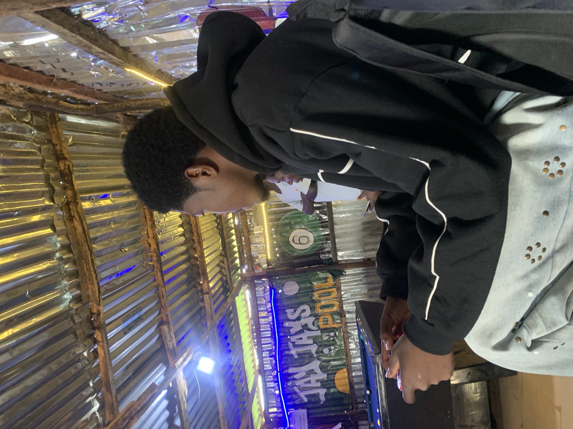 The image depicts an indoor scene, likely a recreational spot, with a corrugated metal roof. In the foreground, a young Black male wearing a black hoodie and light-colored, studded jeans is seated and holding a video game controller, appearing to play a game. Behind him, a pool table is visible, with a banner that reads "JAY JAYS POOL". There are also blue and yellow lights illuminating the area. Another person, wearing a white shirt, is partially visible in the background, facing away from the camera. The environment suggests a casual, possibly local establishment in Durumi, Nigeria.