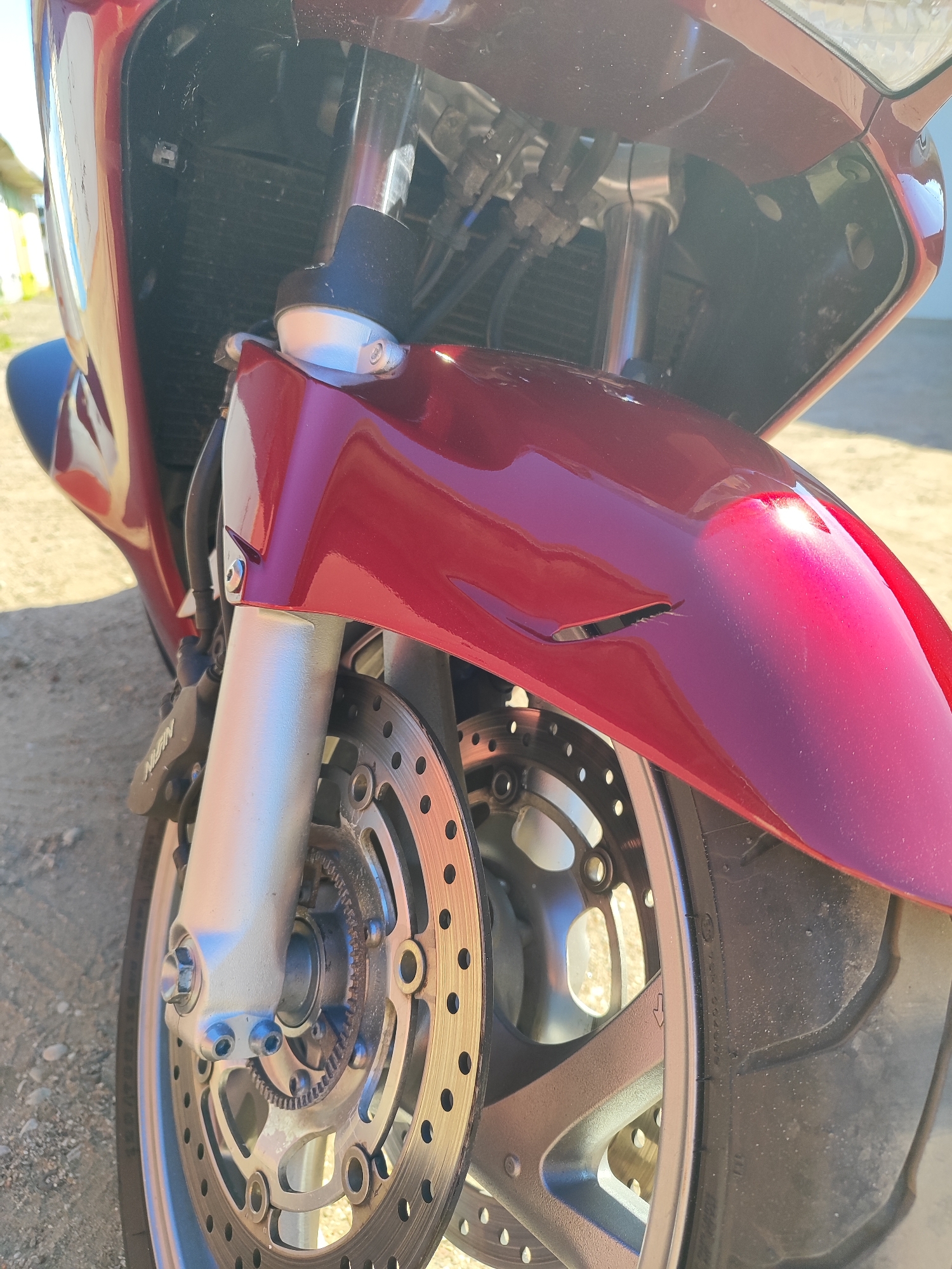 A close-up, low-angle shot shows the front wheel and fender of a dark red motorcycle parked outdoors. The motorcycle is a Honda VFR, based on visible markings. The fender has a noticeable crack and a dent on its right side. The front brake disc, caliper, and suspension fork are clearly visible. The background suggests a rural or suburban setting with gravel and a hint of a building and fence. The image does not depict any people or specific locations in Hrodna, Belarus, beyond the general outdoor environment.