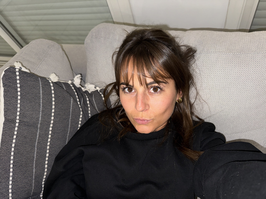 A woman with brown hair and bangs lies on a gray sofa with a striped gray and white pillow. She wears a black sweatshirt and gold earrings. Her expression is neutral, with a slight pout. The lighting is soft, and the background includes part of the ceiling. The image is likely a selfie taken at home.