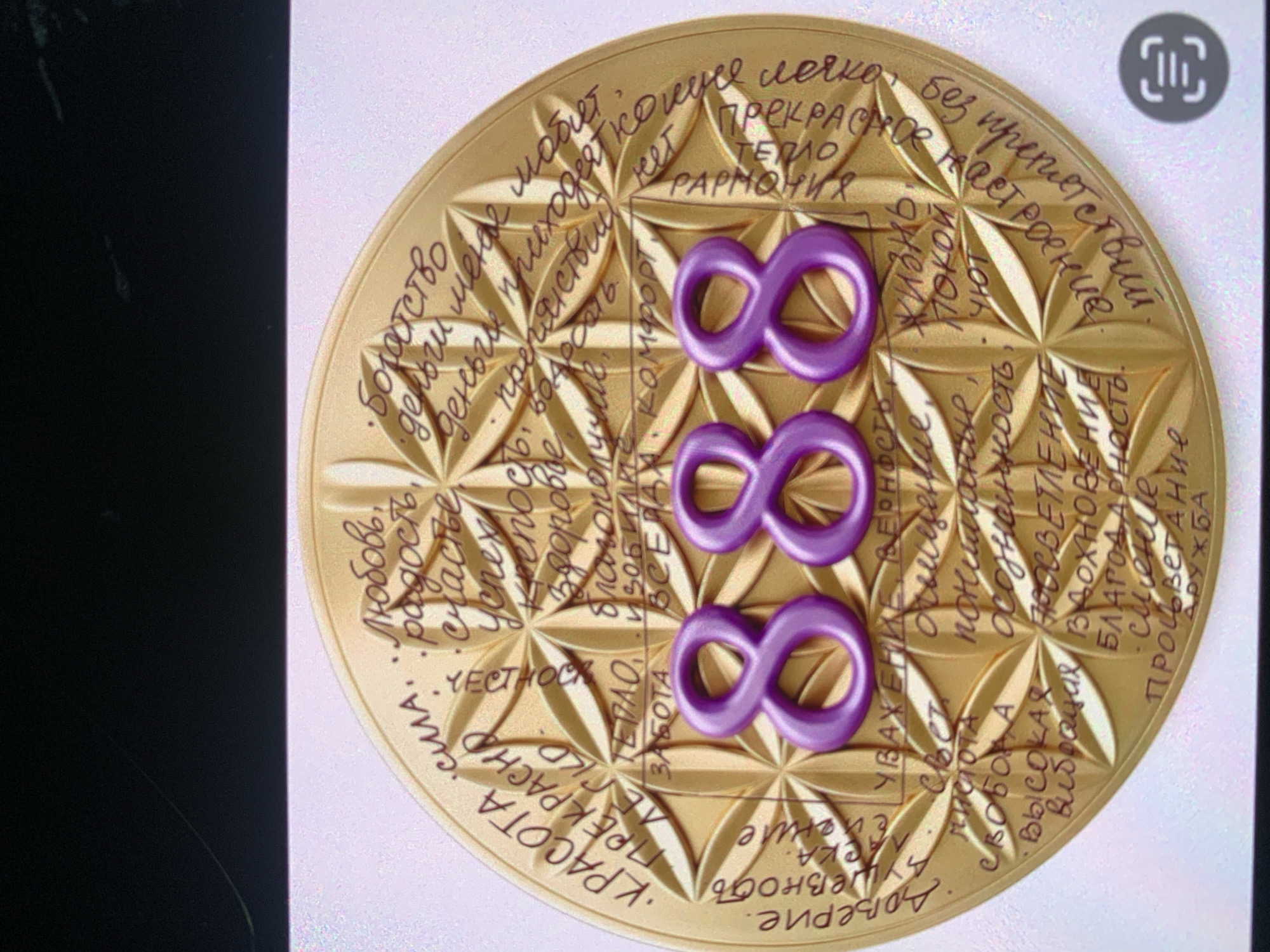 The image shows a close-up, angled shot of a gold-colored, circular amulet or charm. The amulet is adorned with intricate geometric patterns and inscribed with text in Russian. Prominently displayed in the center are three large, raised, purple numbers that spell out "888". The text on the amulet appears to be a list of positive concepts and aspirations, such as "love," "happiness," "wealth," "peace," and "friendship." The amulet is set against a blurred background, and the lighting suggests it might be indoors. There is no discernible location in the city of Sevastopol, Ukraine.